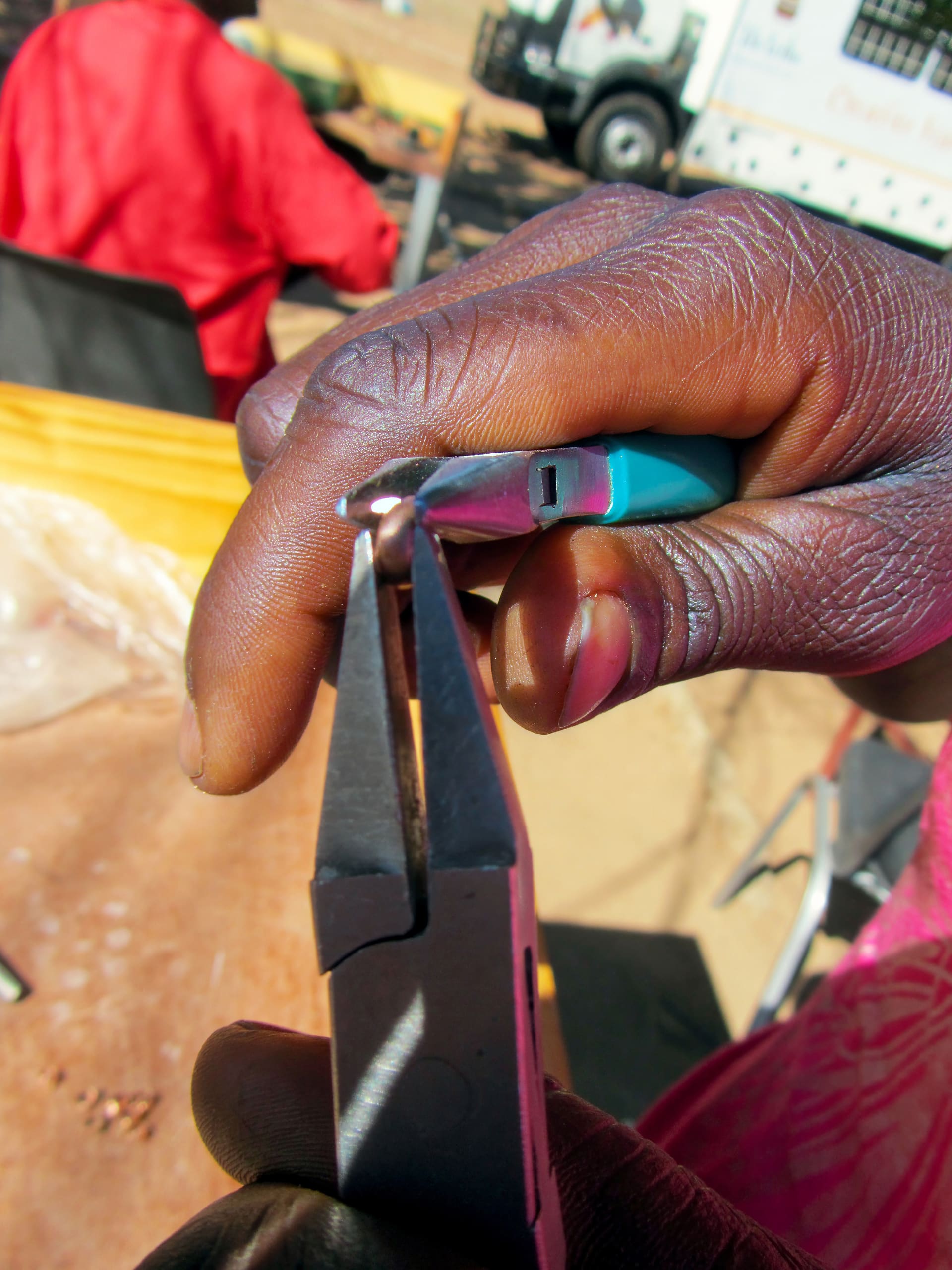 A close-up of a person's hand using pliers to bend a small metal piece. The person's dark skin and textured fingers are visible. The background shows a blurred figure in red and part of a parked vehicle. The focus is on the hands and the tool.