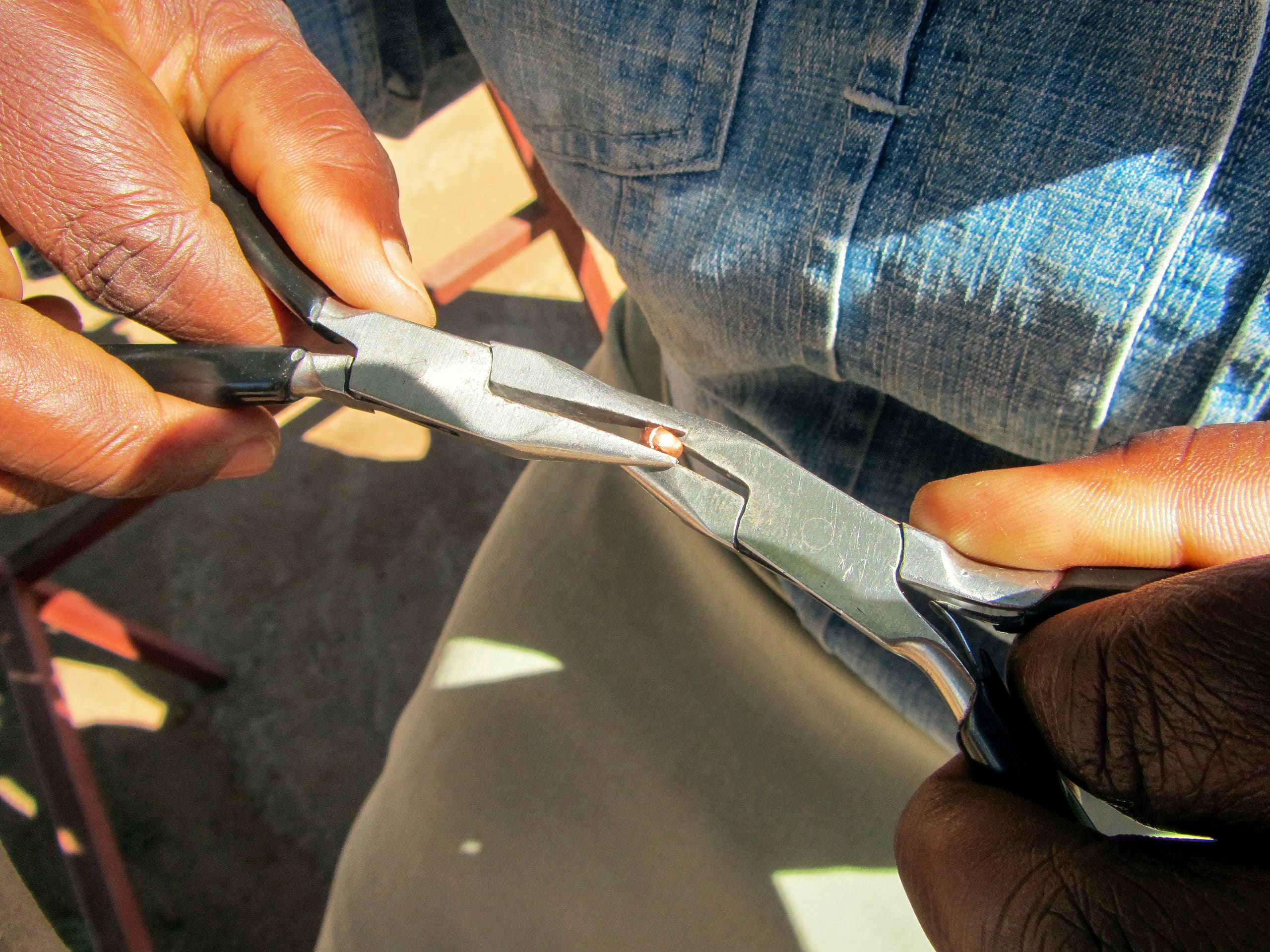 Close-up of hands using two pairs of pliers to bend a small piece of wire. The person is wearing a blue denim shirt and beige trousers, and is seated. The scene is well-lit, showing detail on the tools and hands.
