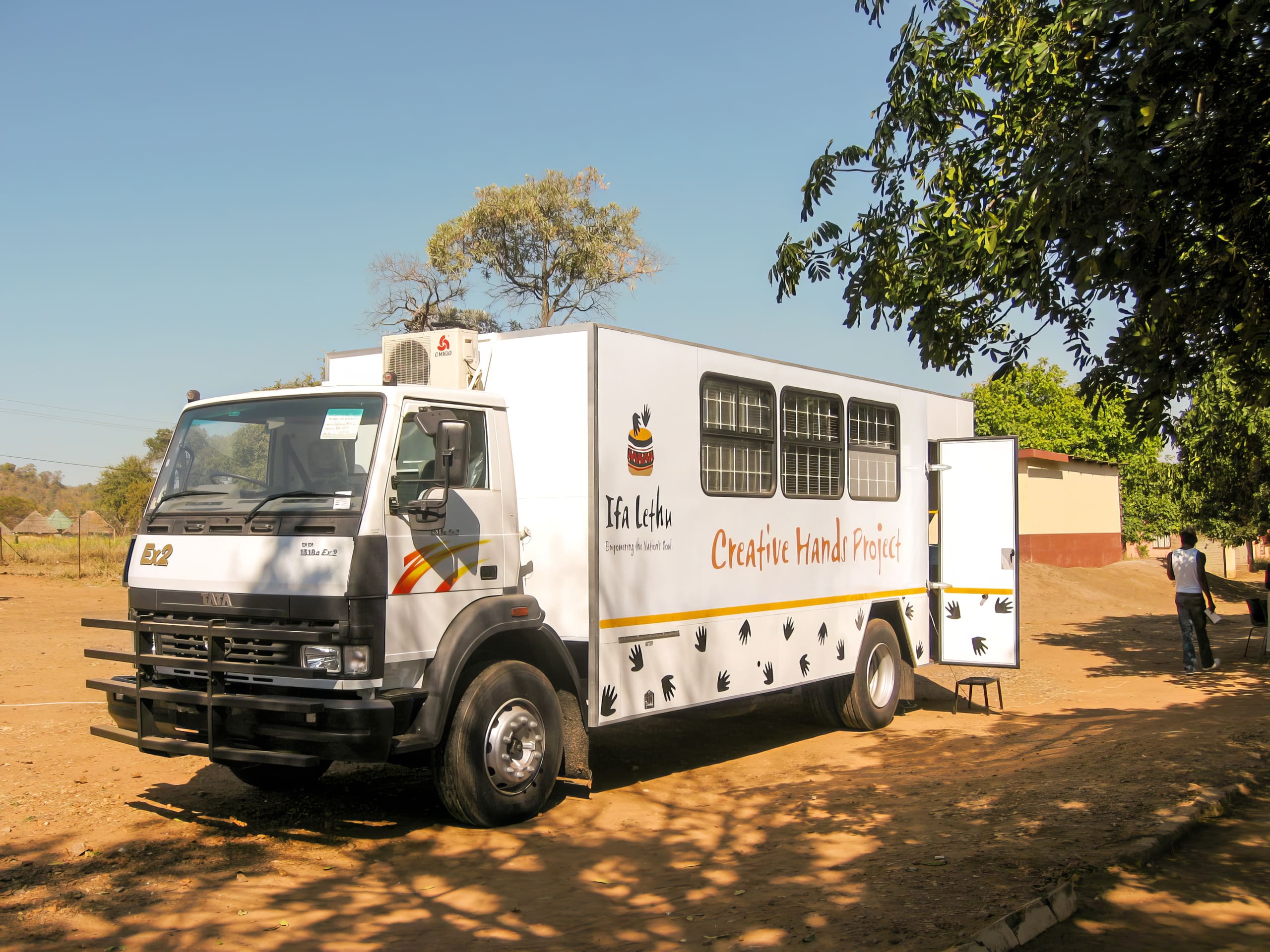 A white mobile lorry labelled “Creative Hand Project” and “Ita Letu” is parked on a dirt road near trees and buildings. The lorry features black handprints and a cat logo. Sunlight casts shadows, and a person stands in the background next to a building.