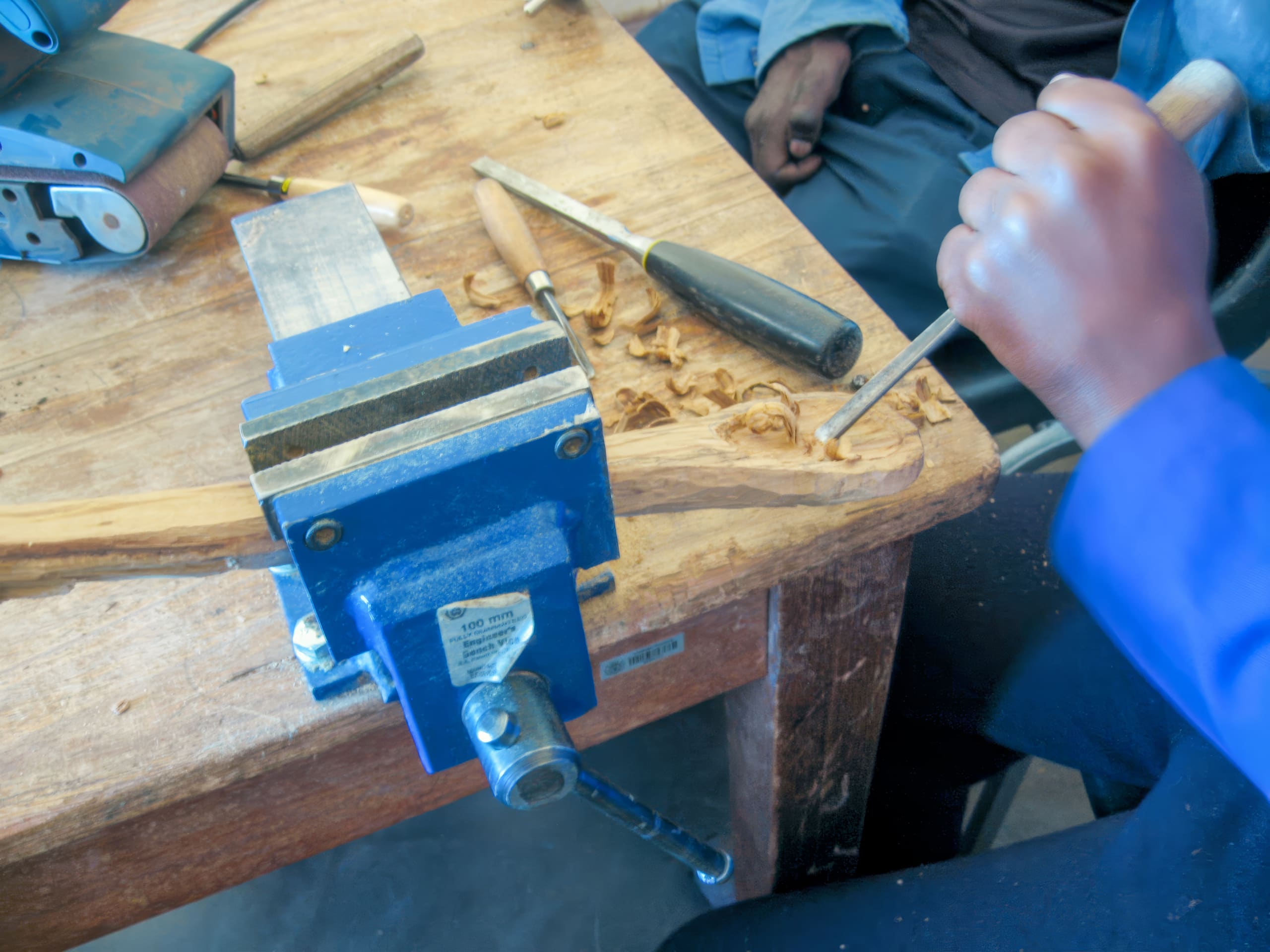 A person uses a chisel and hammer to carve wood held in a blue vice on a workbench, with wood shavings and tools scattered on the wooden table. Only their hand and part of their arm are visible.