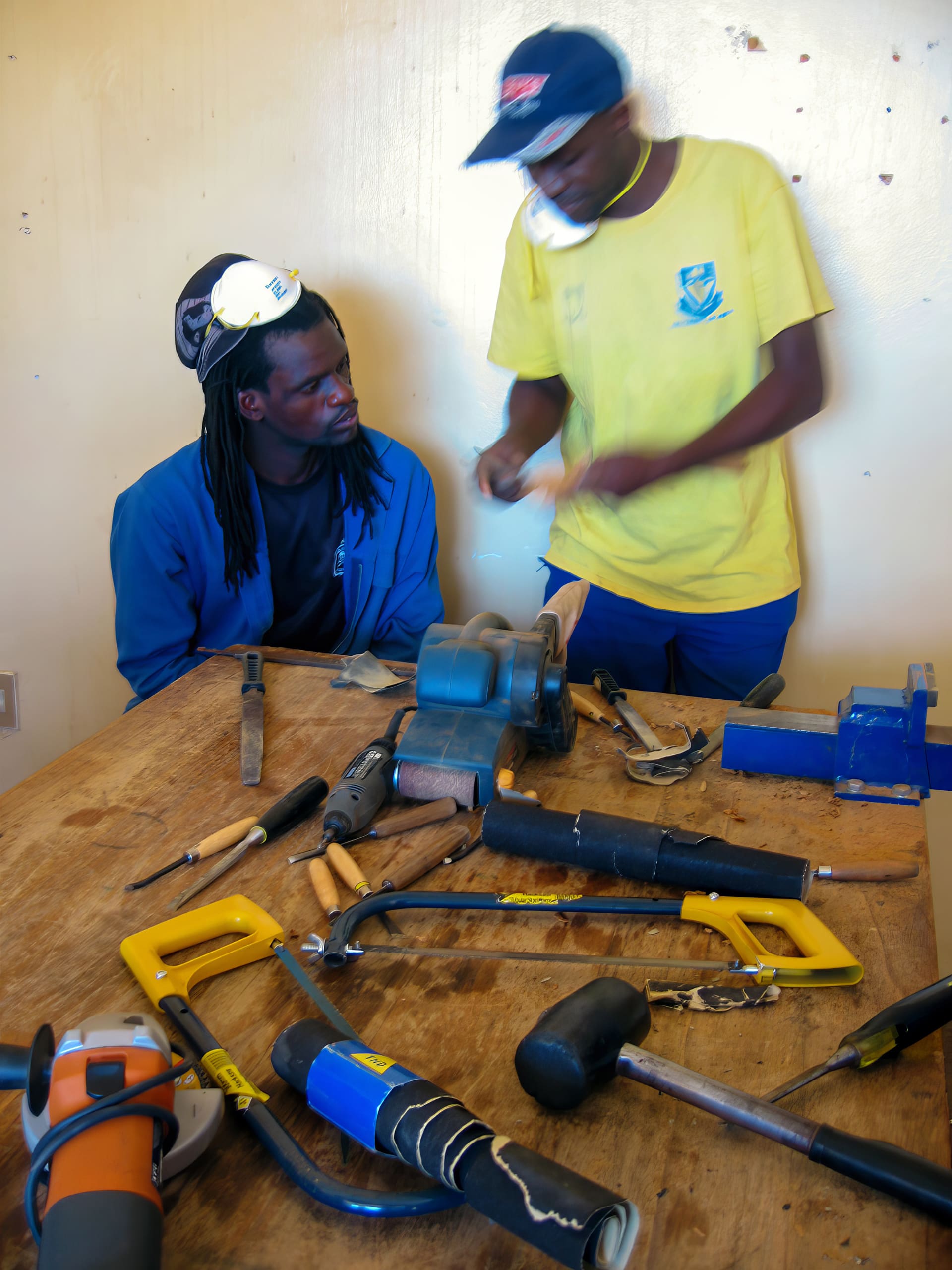 Two men work in a workshop. One, seated, listens intently whilst the standing man explains, gesturing over a table cluttered with hand tools, saws, hammers, and a grinder. The background is plain, and both wear casual clothes; one has a face mask on his head.