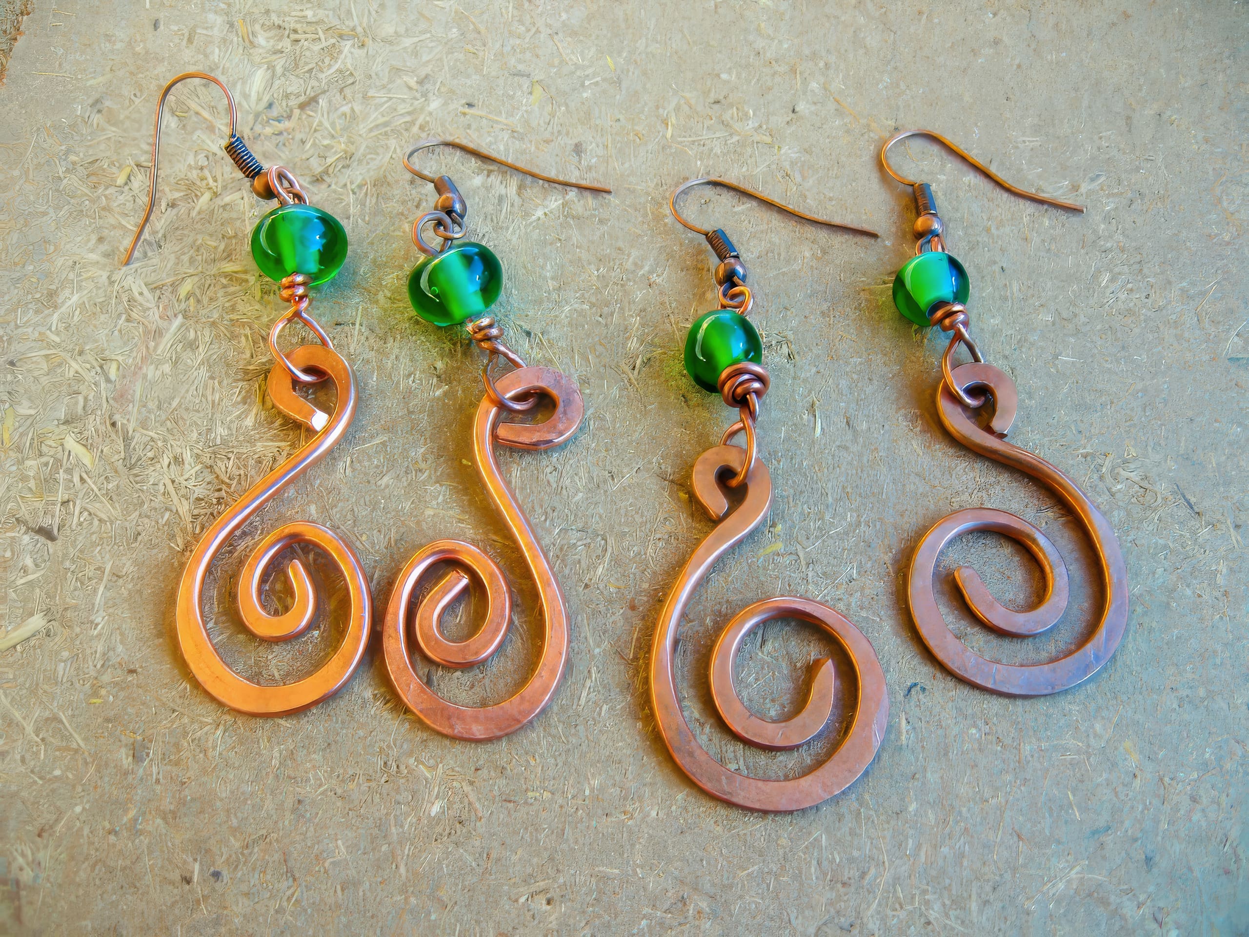Four hand-crafted earrings lay on a textured brown surface. Each earring features a green glass bead above an intricate copper wire swirl design, with two earrings having larger, looser spirals and the other two more tightly wound spirals.