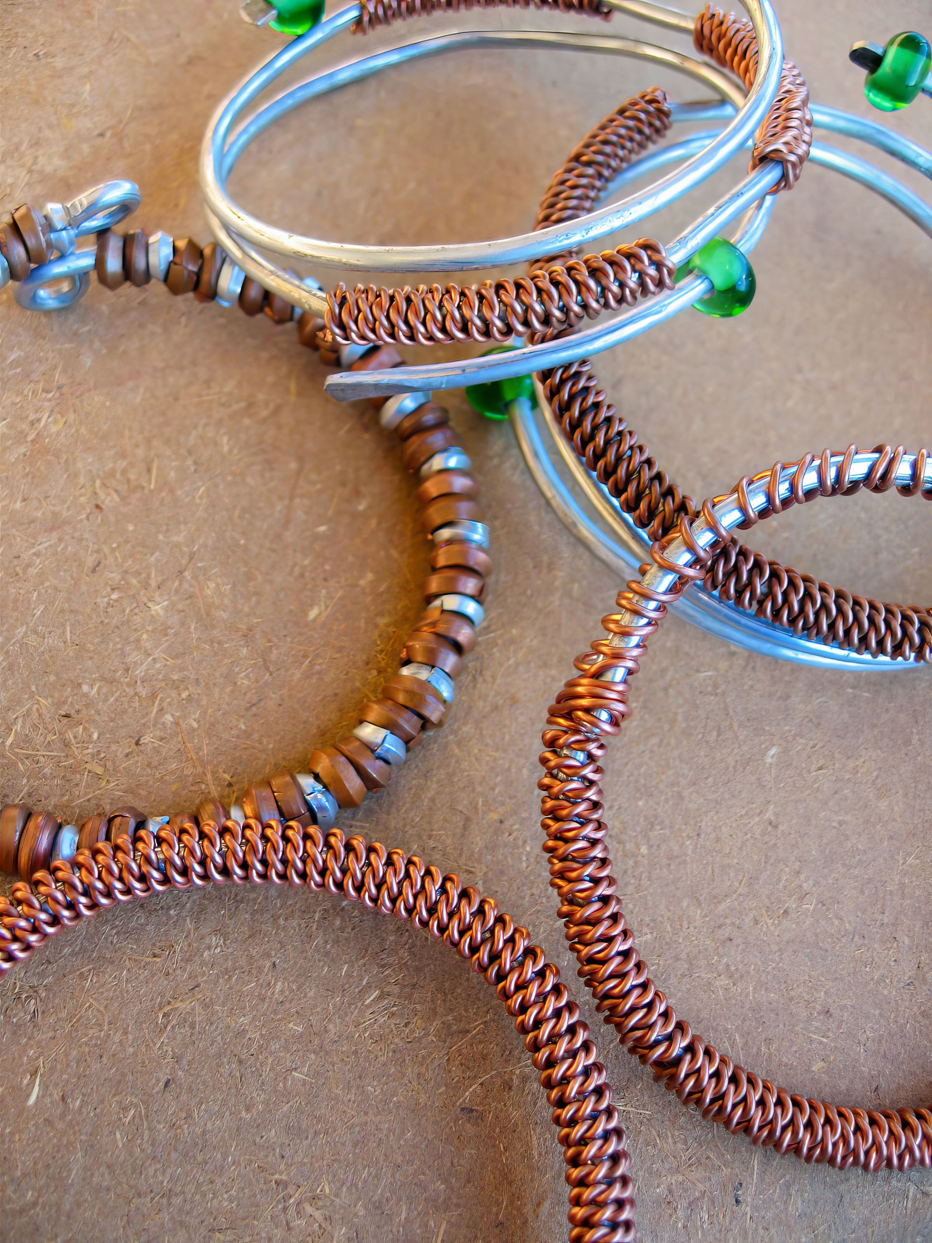 Several handmade wire bracelets rest on a brown surface. Some are copper-coloured with intricate twisted designs, while others are silver with green beads and tan circular beads threaded along the wire. The bracelets overlap and create a textured, artistic arrangement.