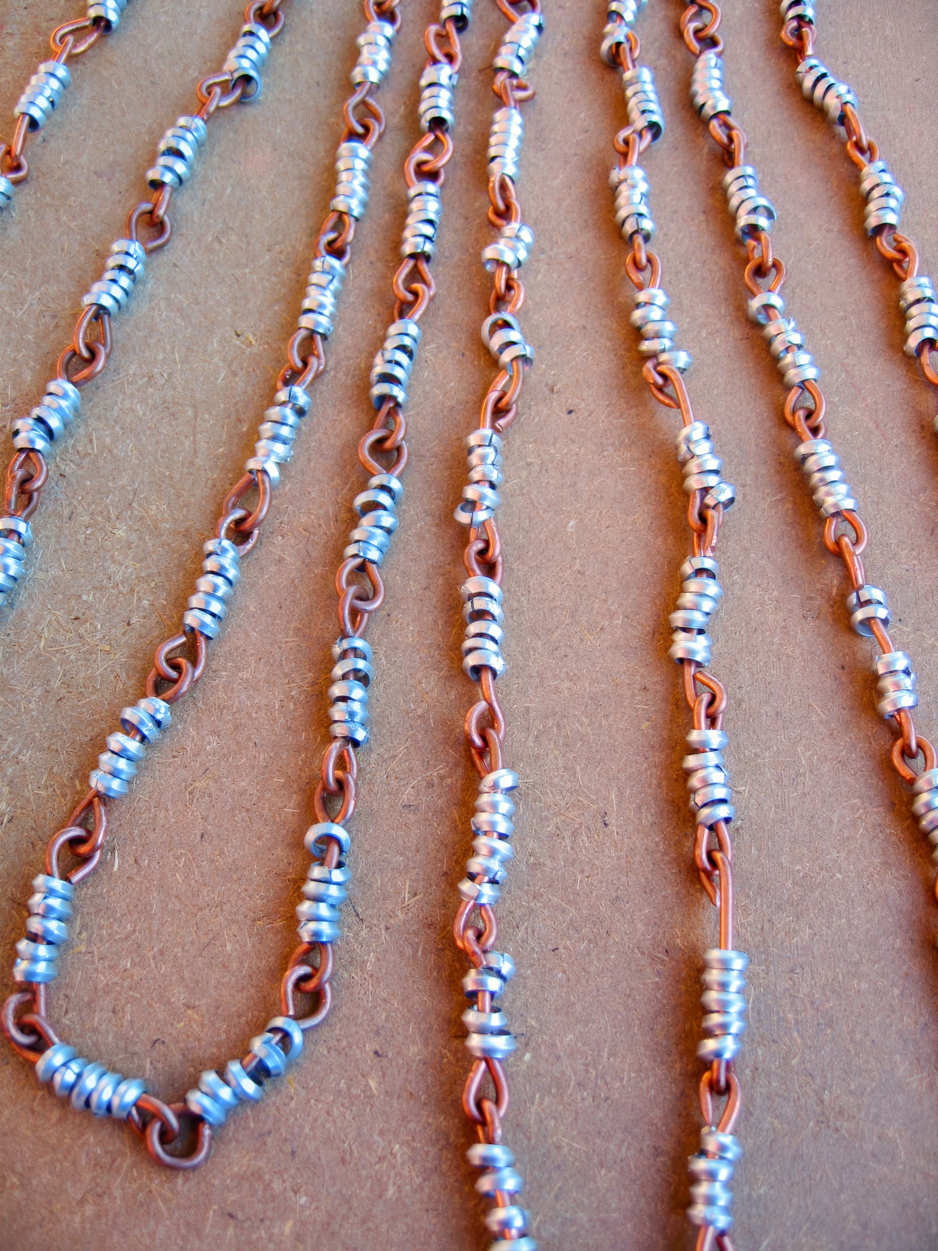 Several strands of copper wire chains lie parallel on a brown surface. Each chain is wrapped with clusters of small, silver-coloured metal rings, creating a beaded effect along the length of the wires. The image is close up and well lit.