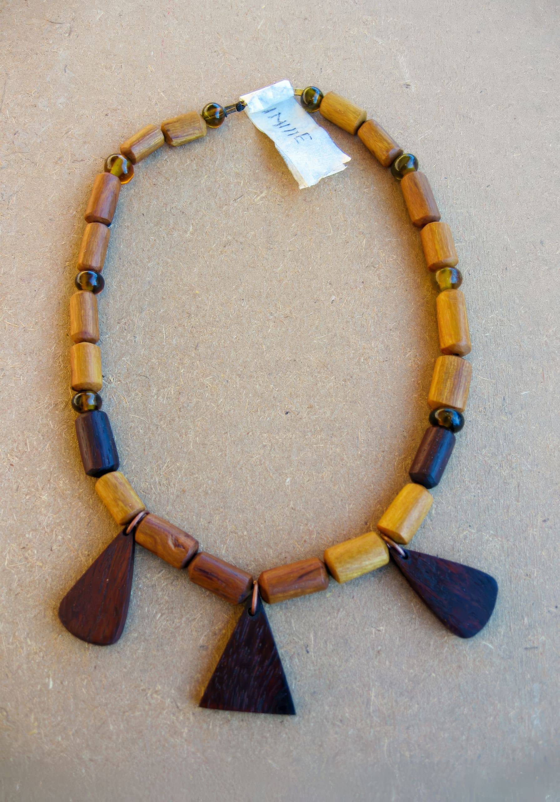A hand-crafted wooden necklace with rectangular light brown beads, small round dark beads, and three large dark wooden pendants in different geometric shapes, laid out in a semicircle on a light brown surface. A white label with 