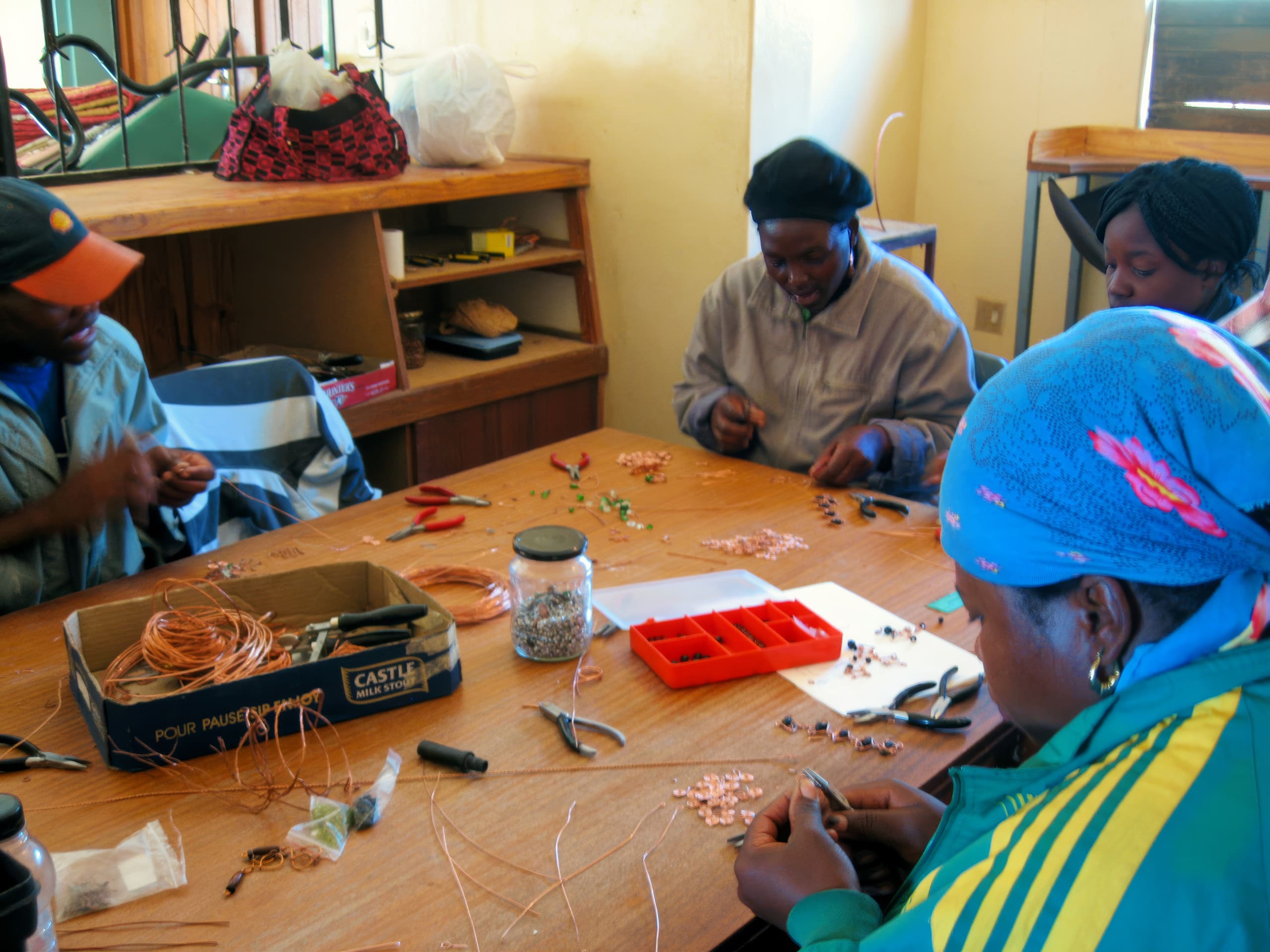 Four people sit around a wooden table making crafts with wire, beads, and tools. A box of copper wire, pliers, and assorted coloured beads are on the table. The room is bright, with shelves and bags in the background. The group appears focused and engaged.