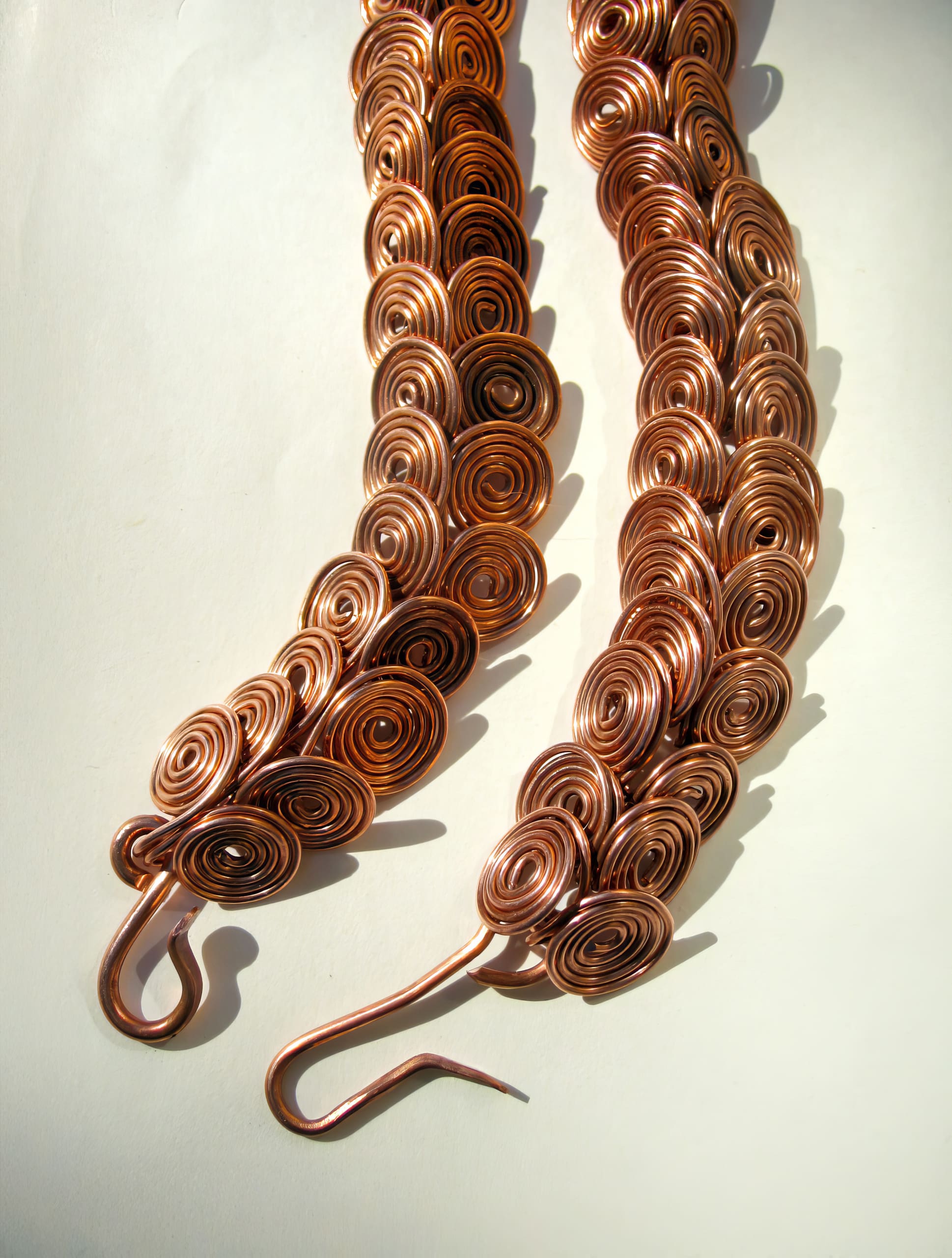 Two intricate copper necklaces made of tightly coiled spirals are arranged side by side on a light surface. Each necklace ends in a hook clasp, and the sunlight highlights the shiny, metallic texture and warm reddish-gold colour of the copper coils.