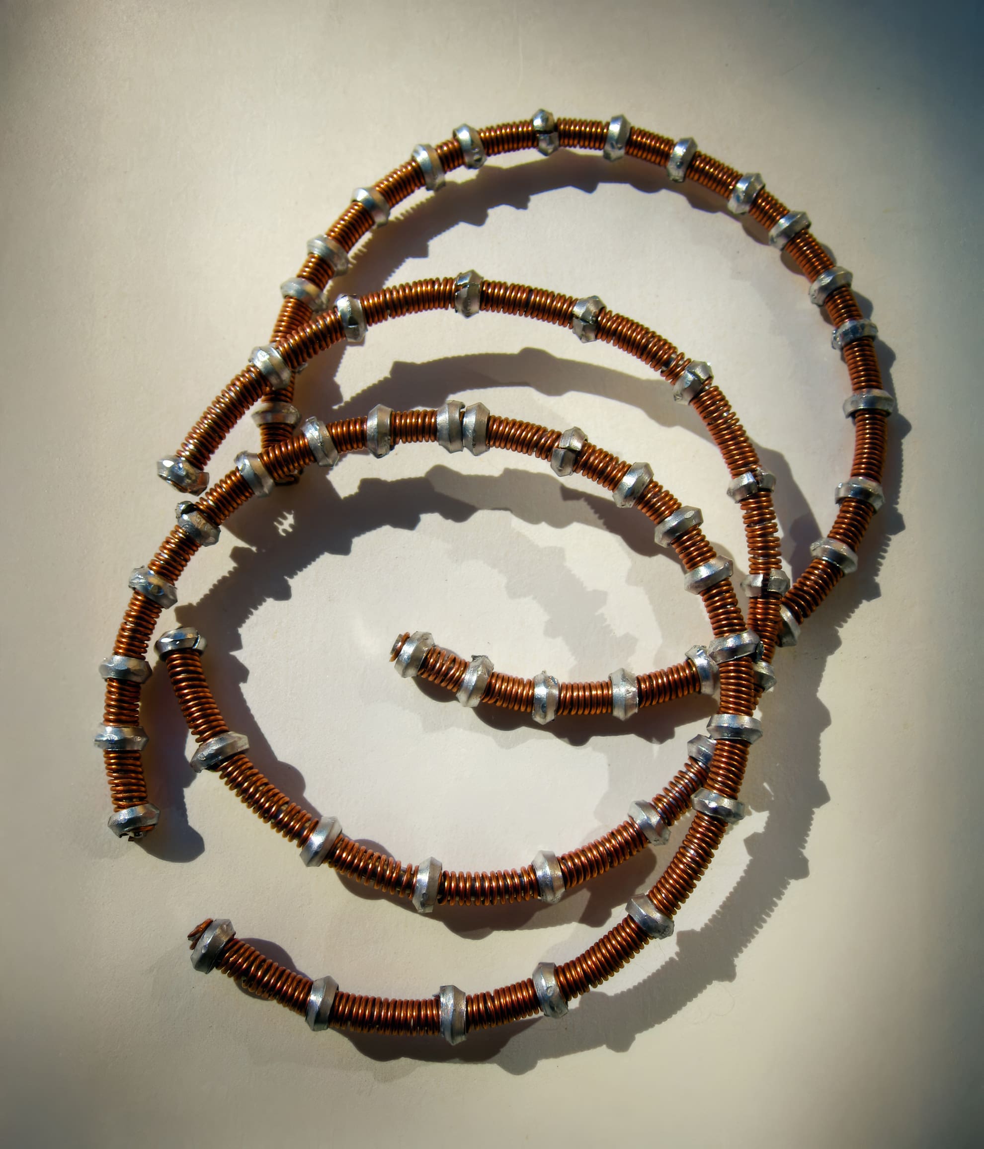A close-up photo of three copper coils arranged in overlapping spirals on a beige surface. The coils are interspersed with evenly spaced silver metal rings, creating a segmented, decorative appearance with prominent shadows.