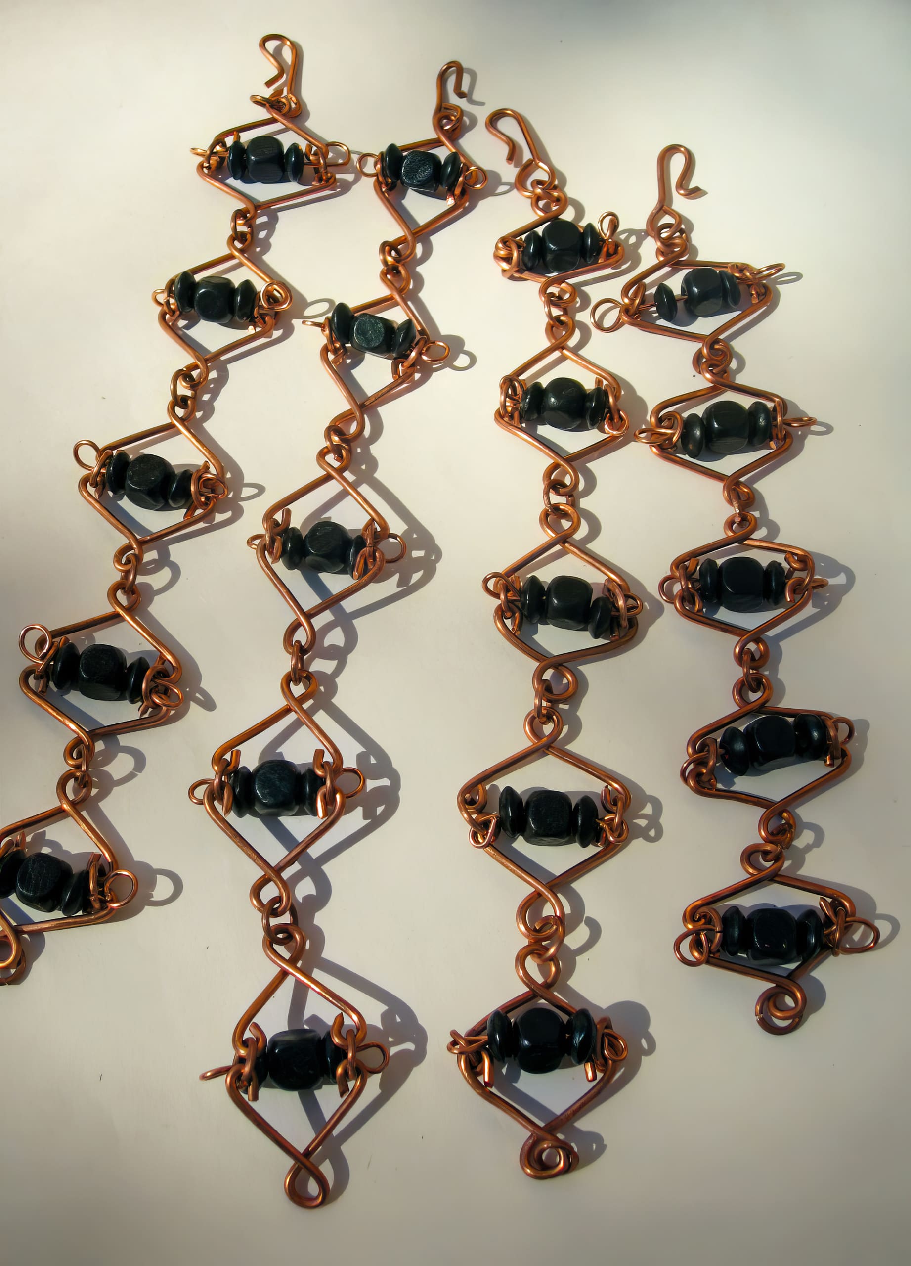 Three strands of copper wire are artistically shaped into geometric diamond patterns, each holding shiny black beads in the centre. Each strand ends with a wire hook, and the design casts intricate shadows on a light background.