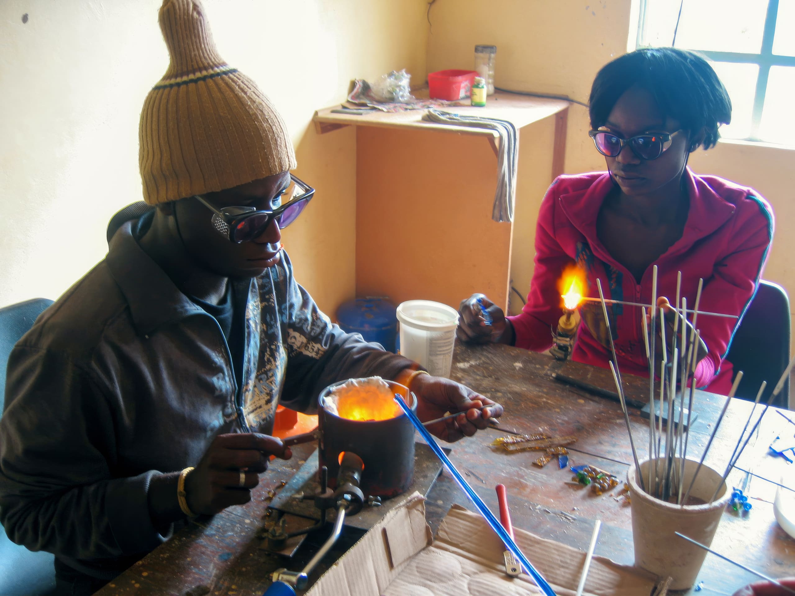 Two people wearing sunglasses work with hot glass at a cluttered table. One uses tools over a small furnace; the other holds a rod with glowing molten glass. The room is sunlit, with shelves, sticks, and glasswork supplies scattered about.