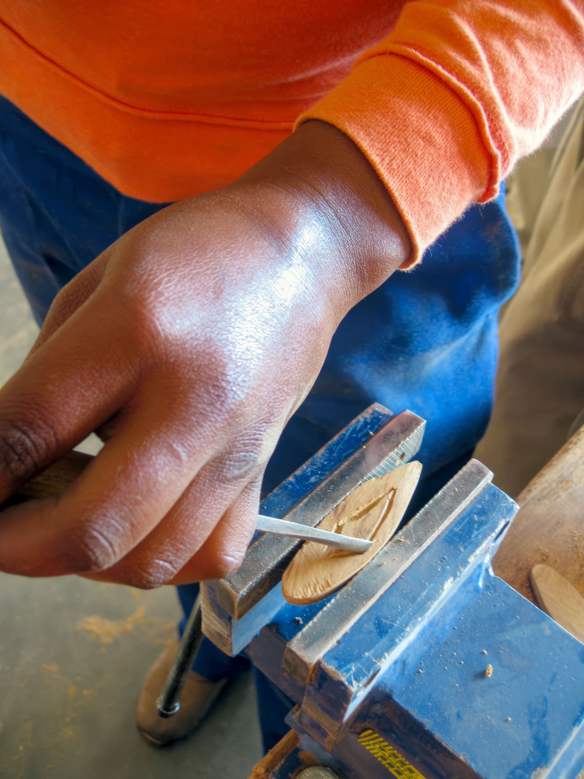 A close-up of a person’s hand in an orange sleeve carving a small wooden piece held in a metal vice. The person uses a chisel or carving tool, wearing blue trousers. The workspace has wood shavings and a smooth, brown tabletop visible.