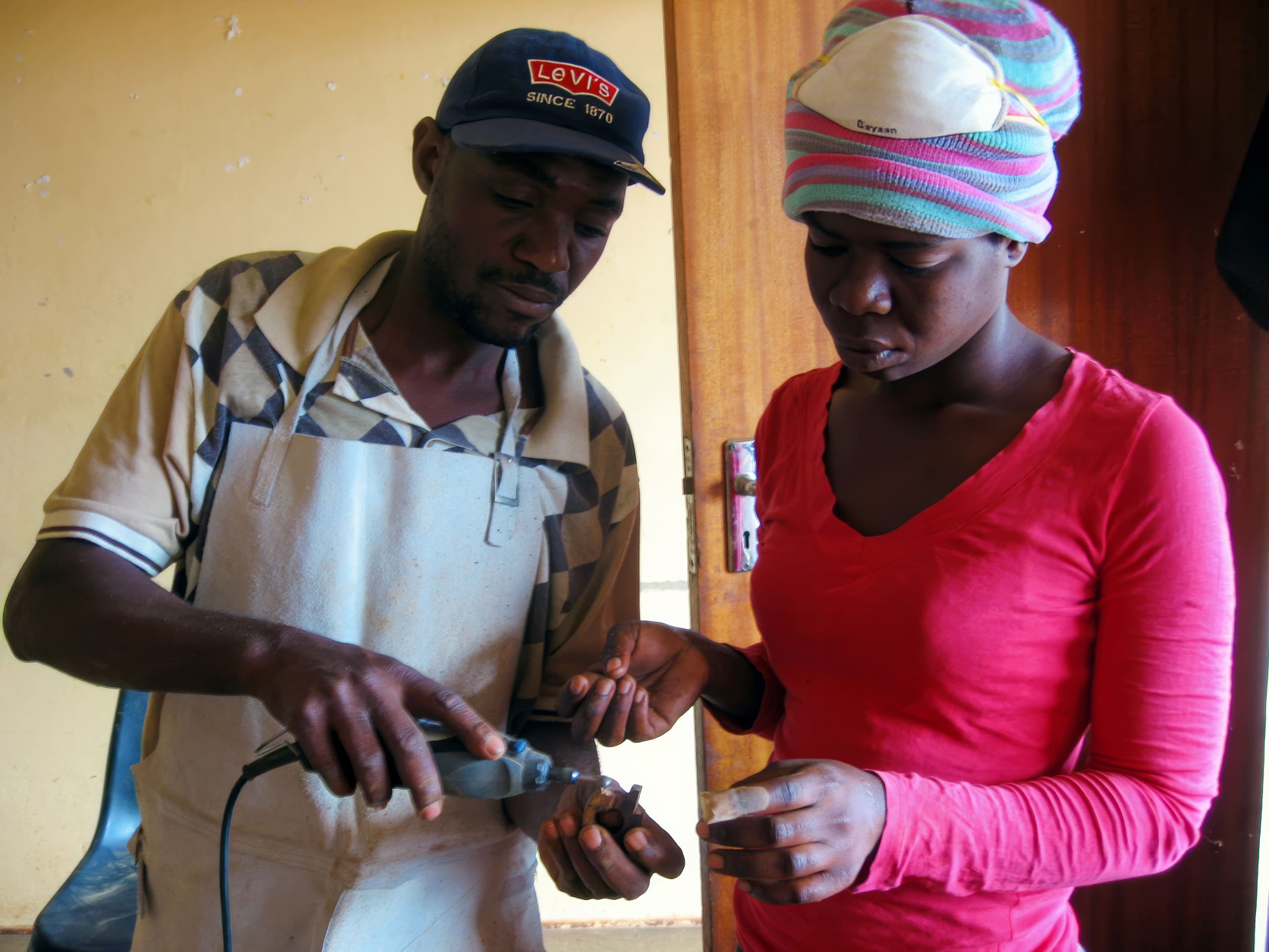 A man wearing a Levi’s cap and apron uses a handheld tool on a woman’s hand. The woman, in a red shirt and striped headscarf, looks down at her hand with a serious expression. They are standing indoors near a wooden door.