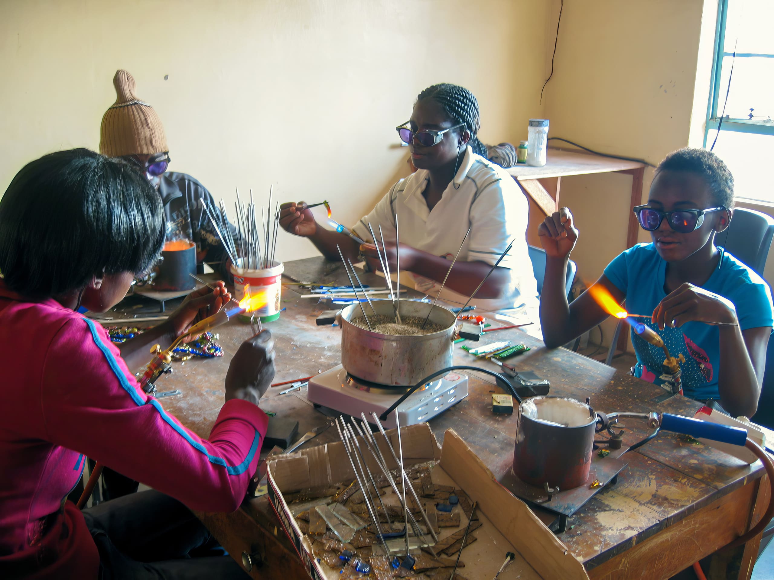 Four people sit around a wooden table making glass art. They wear protective spectacles and use tools to shape glass rods in flames. The table is cluttered with equipment and colourful materials. Sunlight enters from a window on the right, illuminating the workspace.