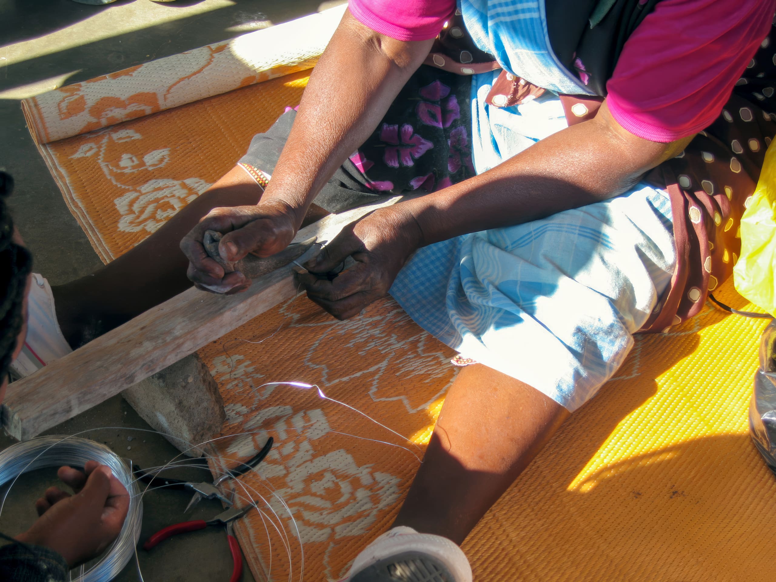 A person sits on an orange patterned mat, wearing a blue skirt and pink shirt, using pliers and a wooden block to shape thin wire. Additional wire and tools are visible nearby. Sunlight casts shadows across the scene.