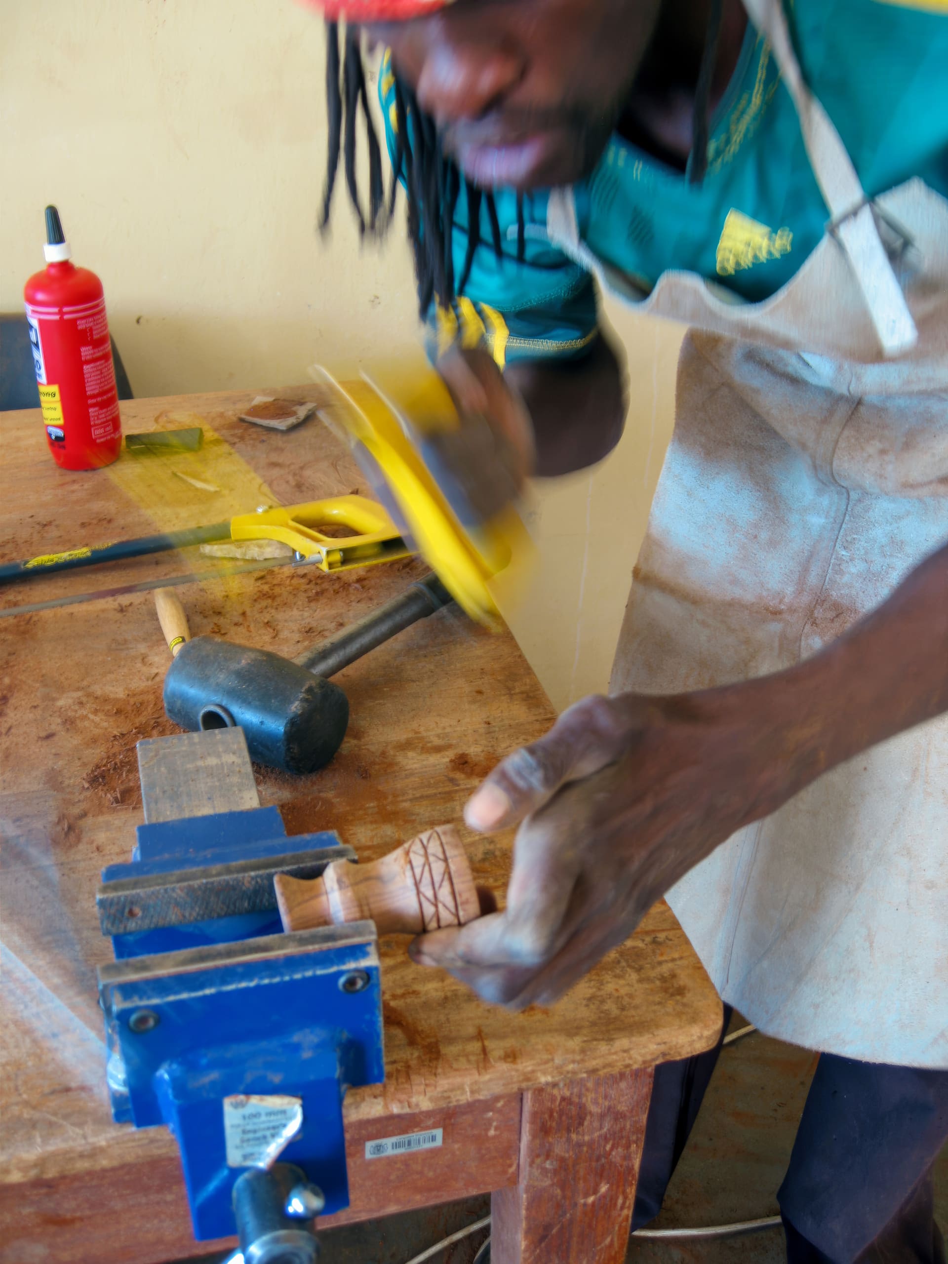 A person wearing a green shirt and apron uses a yellow saw to cut a small piece of wood clamped in a blue vice on a workbench. The table holds tools including a mallet, sanding block, and a red bottle of glue. The scene suggests woodworking activity.
