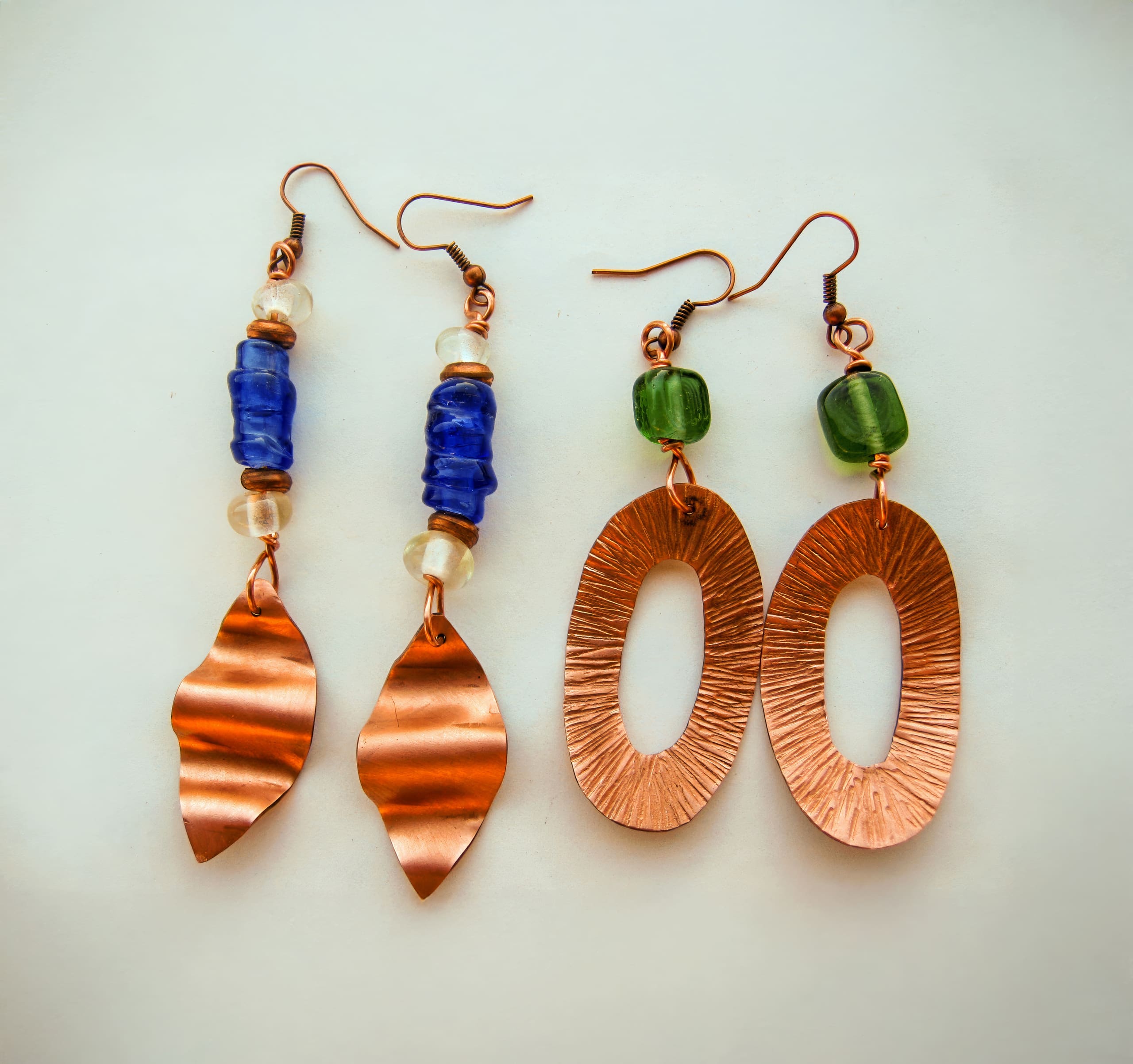 Two pairs of handmade copper earrings: one with wavy copper leaves, blue and white beads; the other with large textured copper ovals and green glass beads. Both pairs have copper hooks and are photographed on a light background.