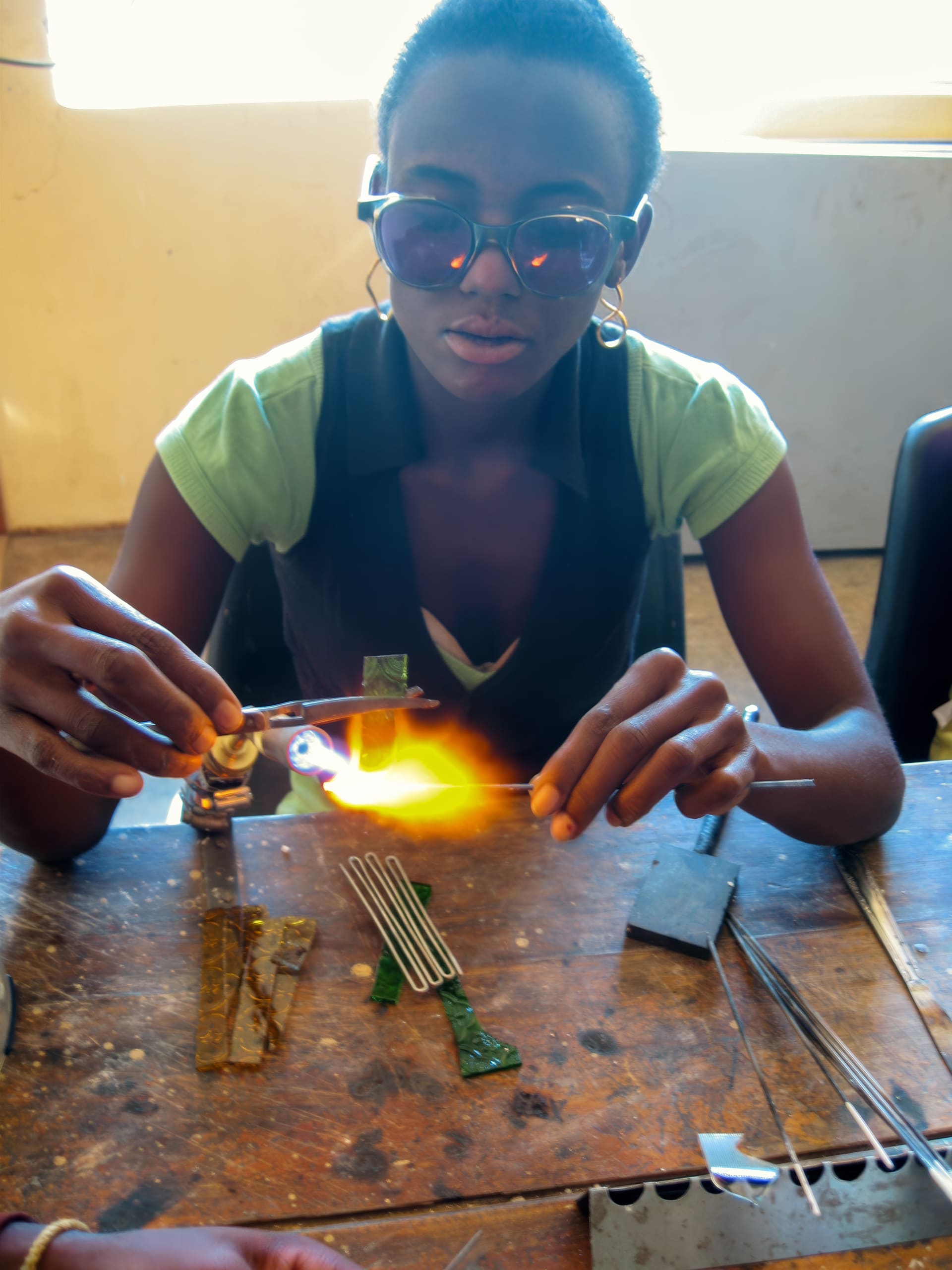 A person wearing dark safety glasses and a green shirt uses a small blowtorch to shape glass on a wooden table, surrounded by glass rods and tools, with a bright flame illuminating their hands and workspace.