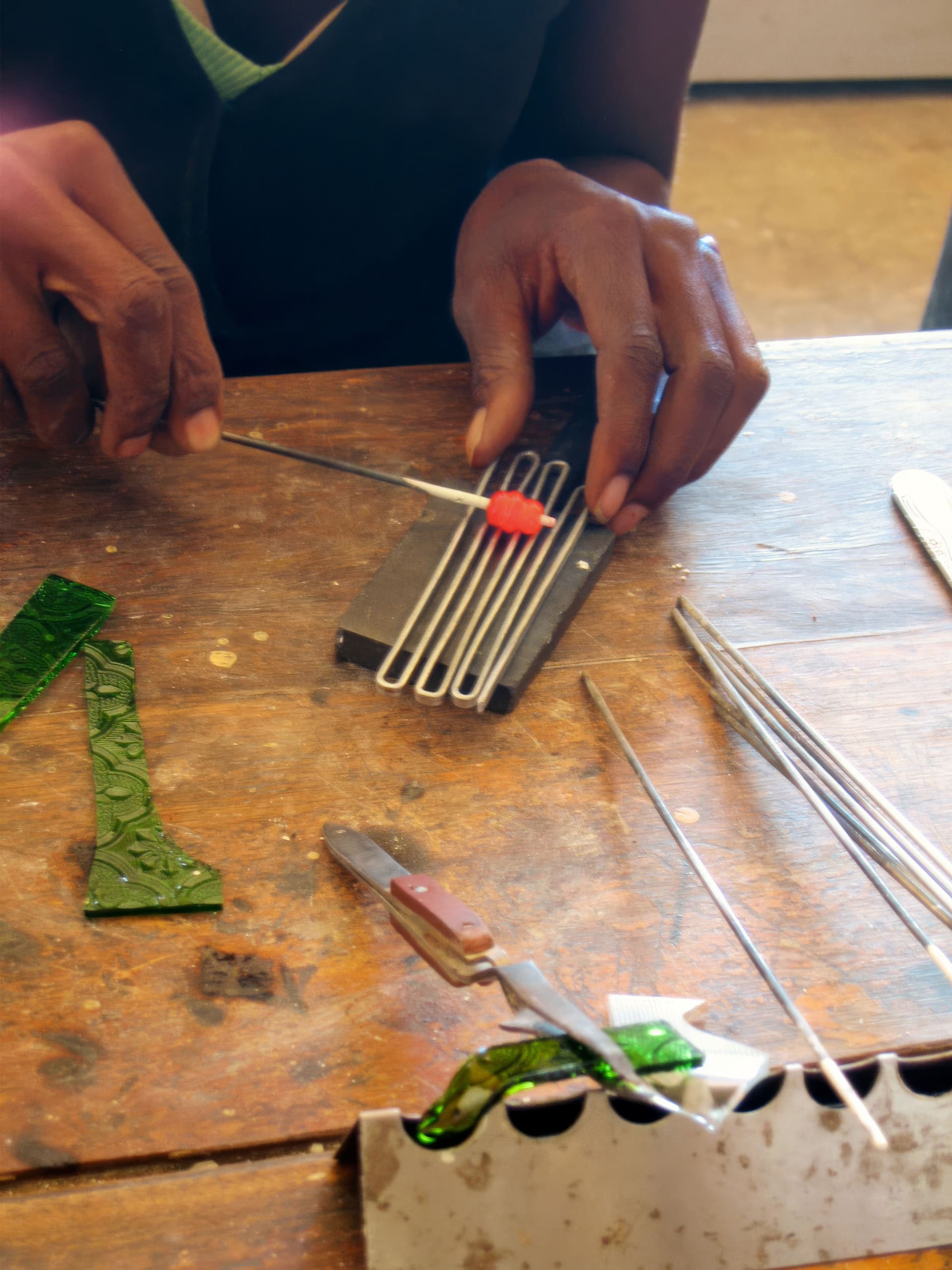 A person uses metal tools to arrange glass rods and a small red glass piece on a work surface. Green glass shards, a knife, and additional rods are scattered on the wooden table, suggesting glasswork or jewellery making in progress.