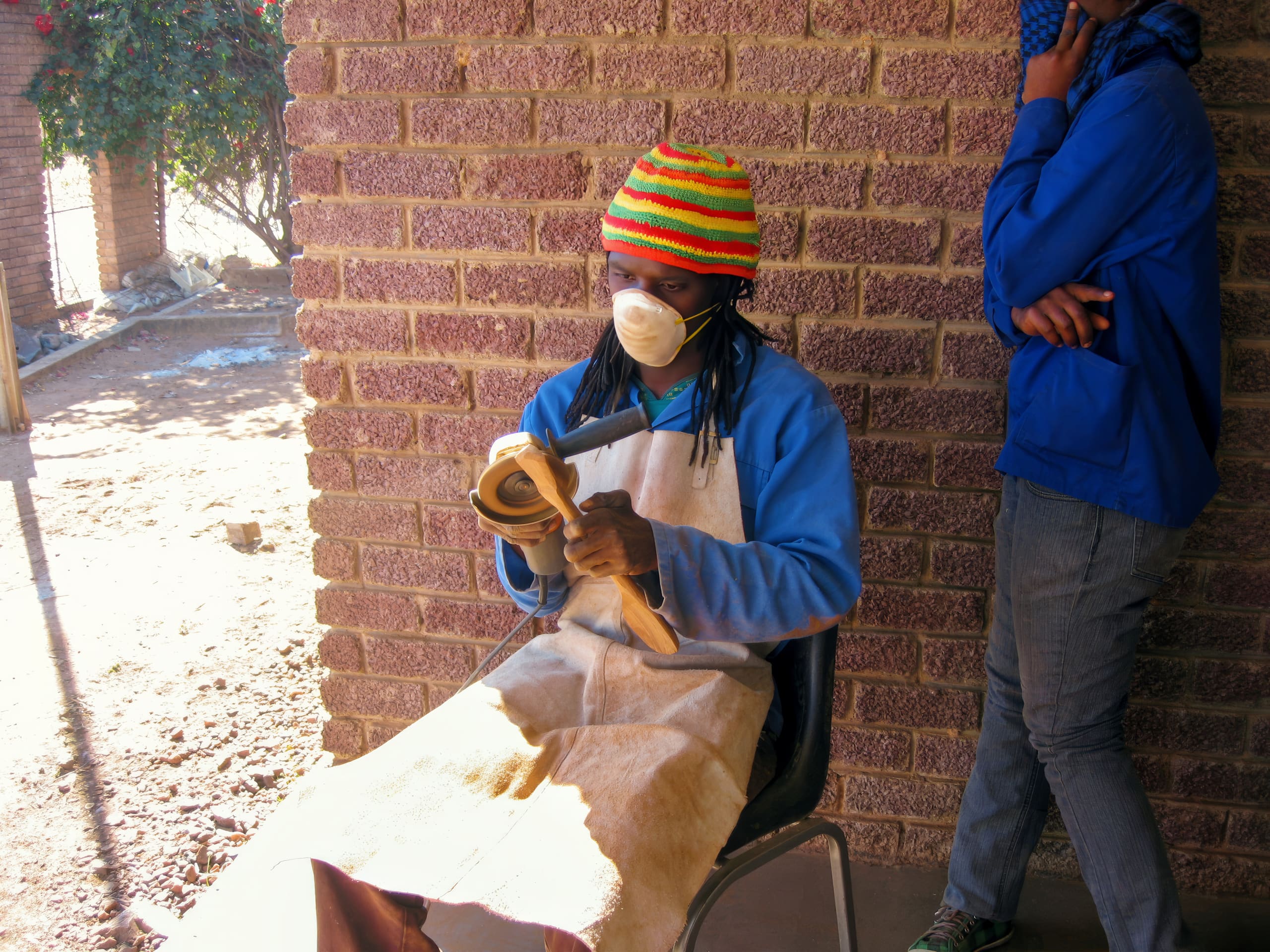 A person wearing a colourful hat, face mask, blue shirt, and apron sits on a chair, carving a wooden object. Another person in blue stands nearby, leaning against a brick wall with arms crossed, partially covering their face with their hand and scarf.
