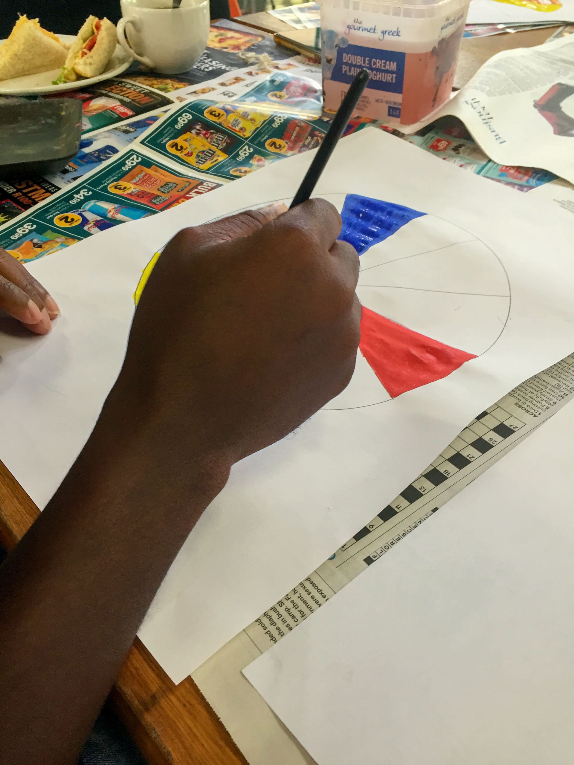 A person’s hand colours a pie chart on white paper using paint and a brush. The chart has red, blue, and yellow sections. Newspapers, a yoghurt tub, food, and other art supplies are scattered on the wooden table.