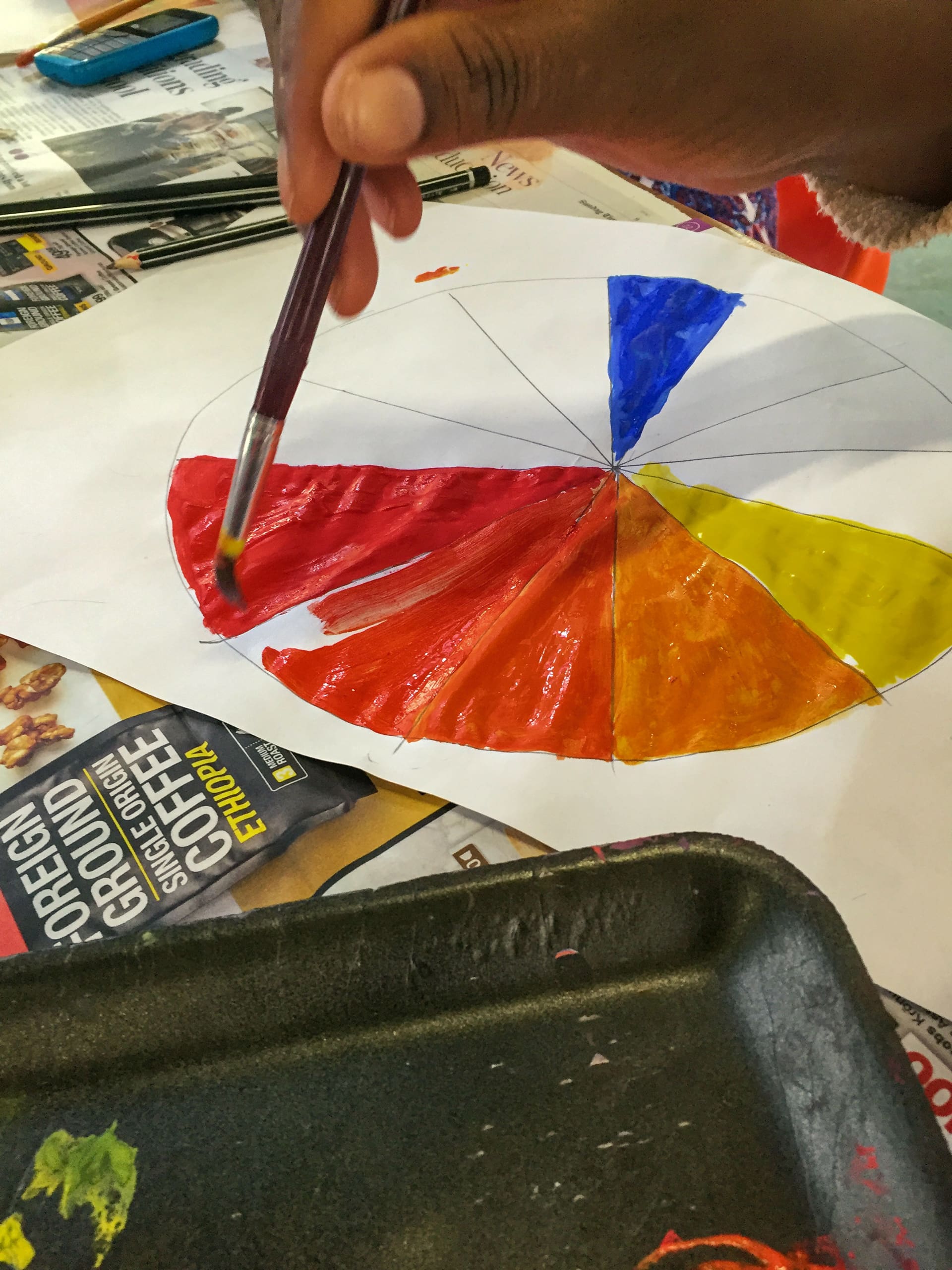 A hand holds a paintbrush, painting sections of a colour wheel on white paper. The wheel includes red, yellow, blue, and orange. Nearby are newspapers, a black tray with paint, and a coffee advertisement on the table.