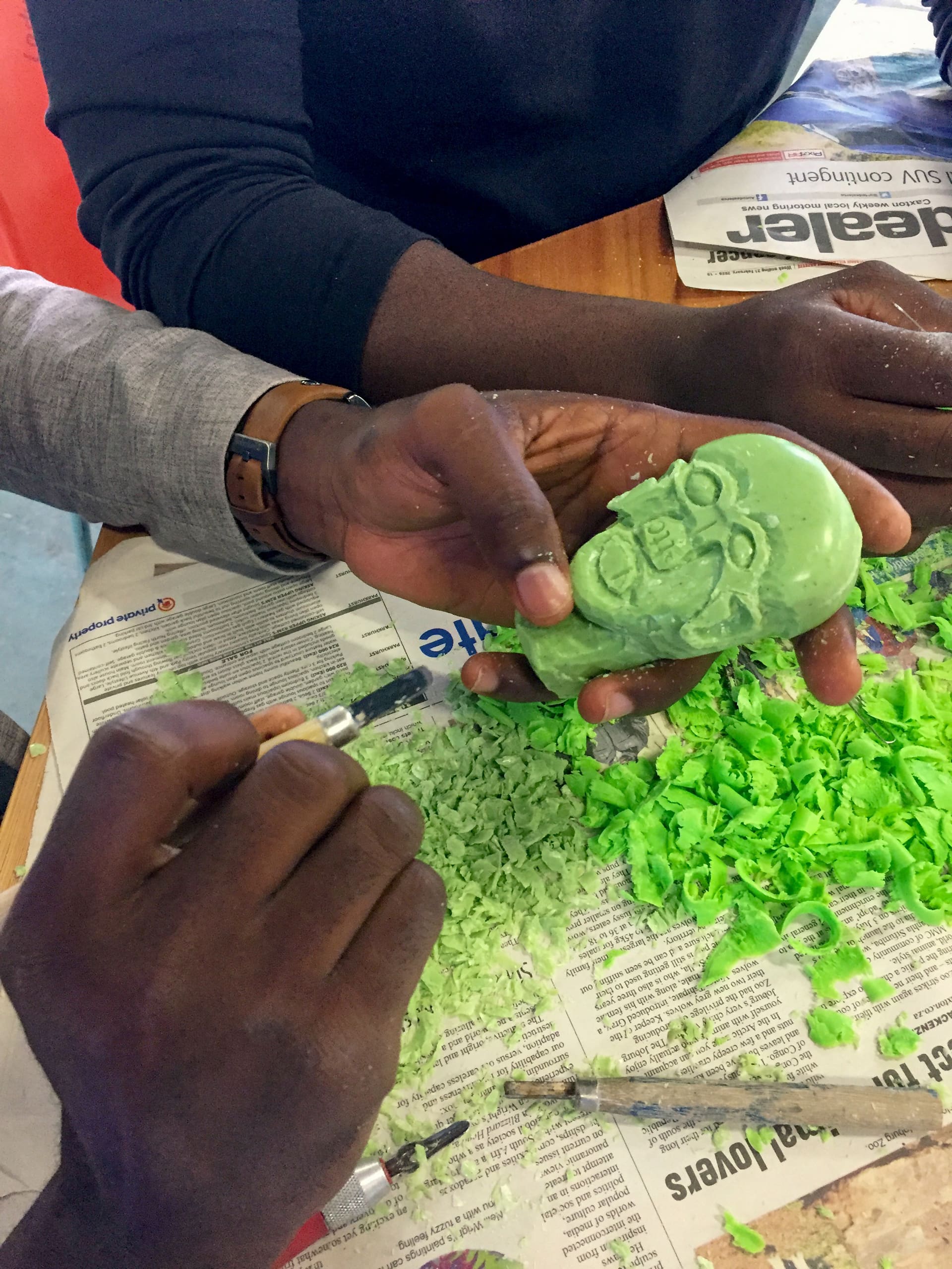 Two hands carve a green bar of soap into the shape of a face, surrounded by soap shavings on a table covered with newspaper. One hand holds the soap, while the other uses a carving tool to add detail to the sculpture.