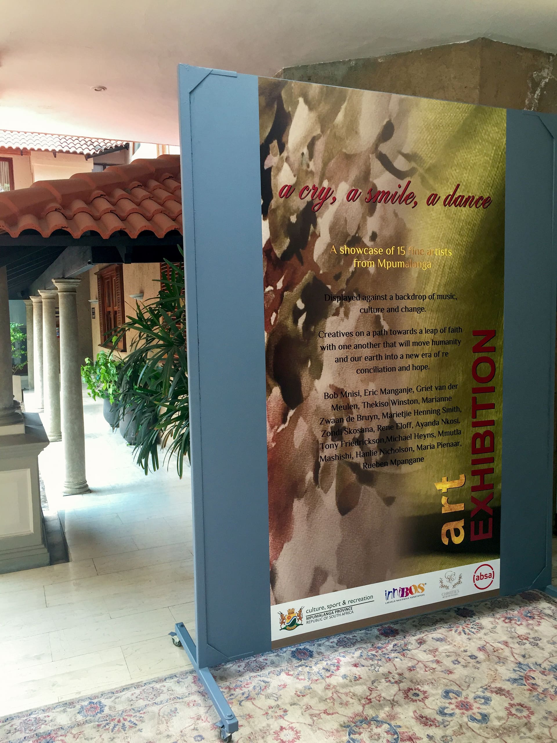 A large vertical sign stands indoors near columns and plants. The sign reads: 