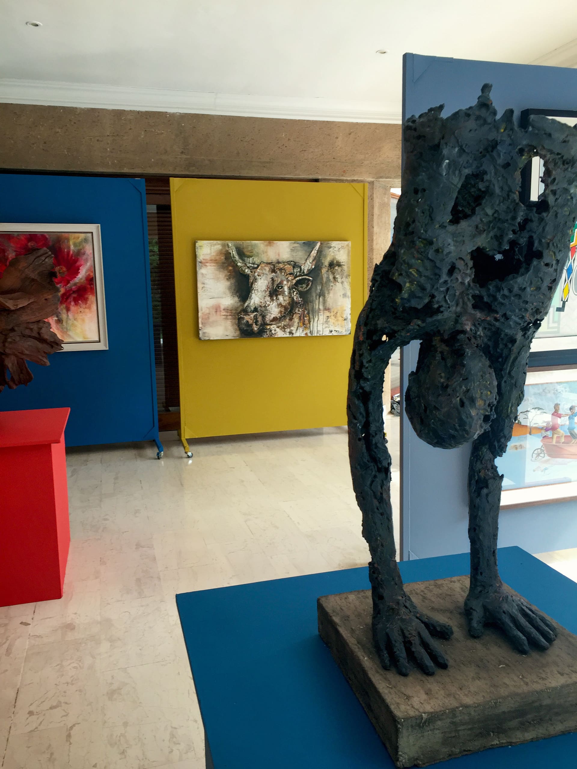 A modern art gallery displays abstract sculptures and paintings. In the foreground, a dark metal sculpture of a figure bends forward on a blue plinth. Behind it, colourful paintings hang on blue and yellow walls, including art of an owl and textured abstract forms.