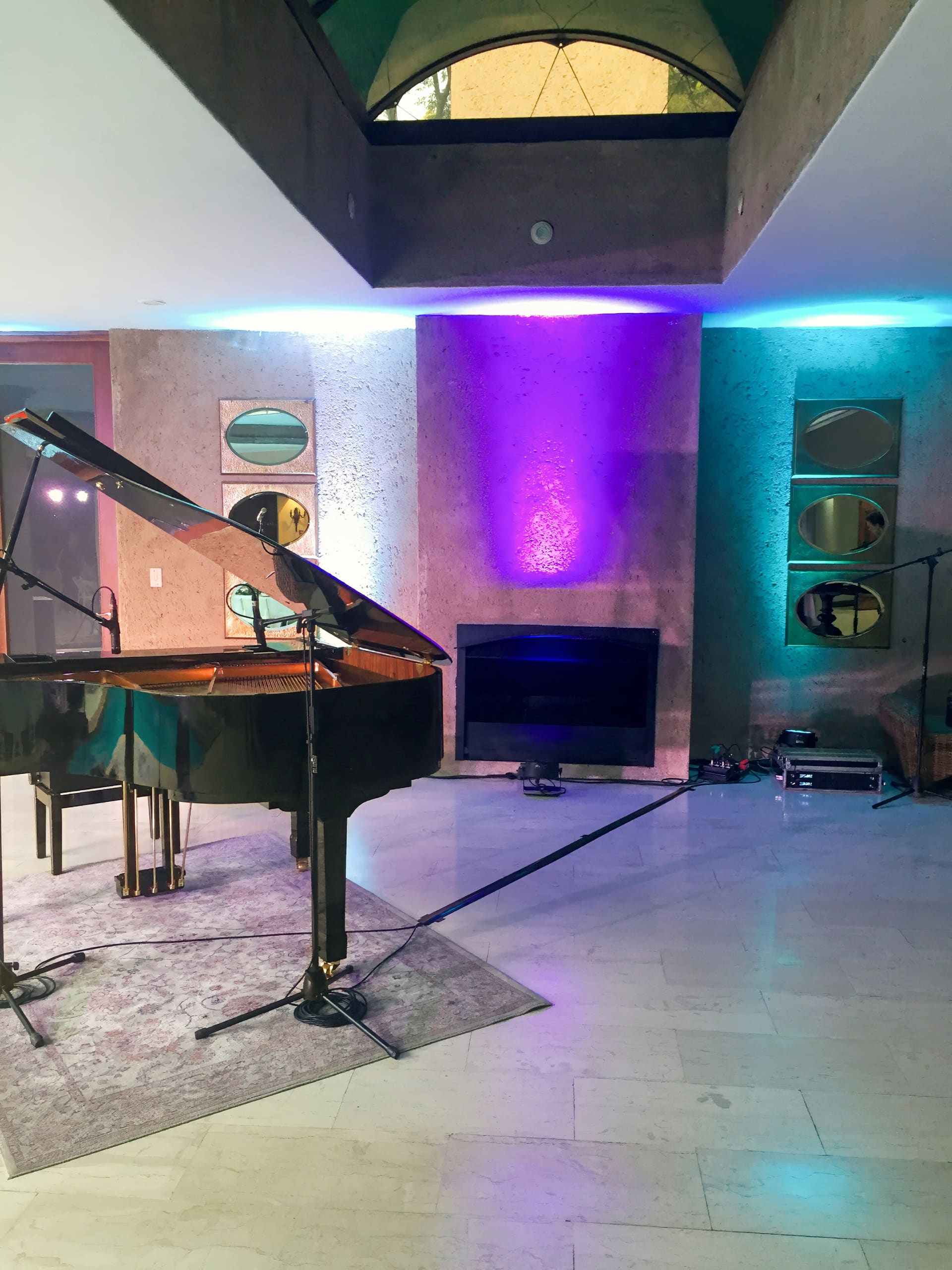 A grand piano is set up in a modern room with textured walls, circular mirrors, colourful LED lighting (purple and blue), and audio equipment, including microphones on stands, near a black fireplace and a high ceiling with an upper window.