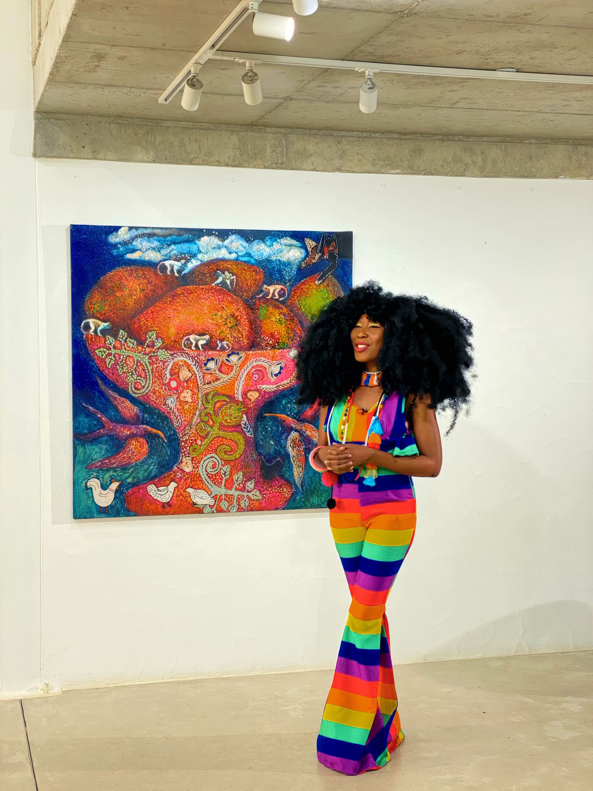 A woman with voluminous curly black hair, wearing a vibrant rainbow-striped jumpsuit, stands smiling in front of a colourful, abstract elephant painting displayed on a white gallery wall. She clasps her hands and looks relaxed amid an art exhibition.