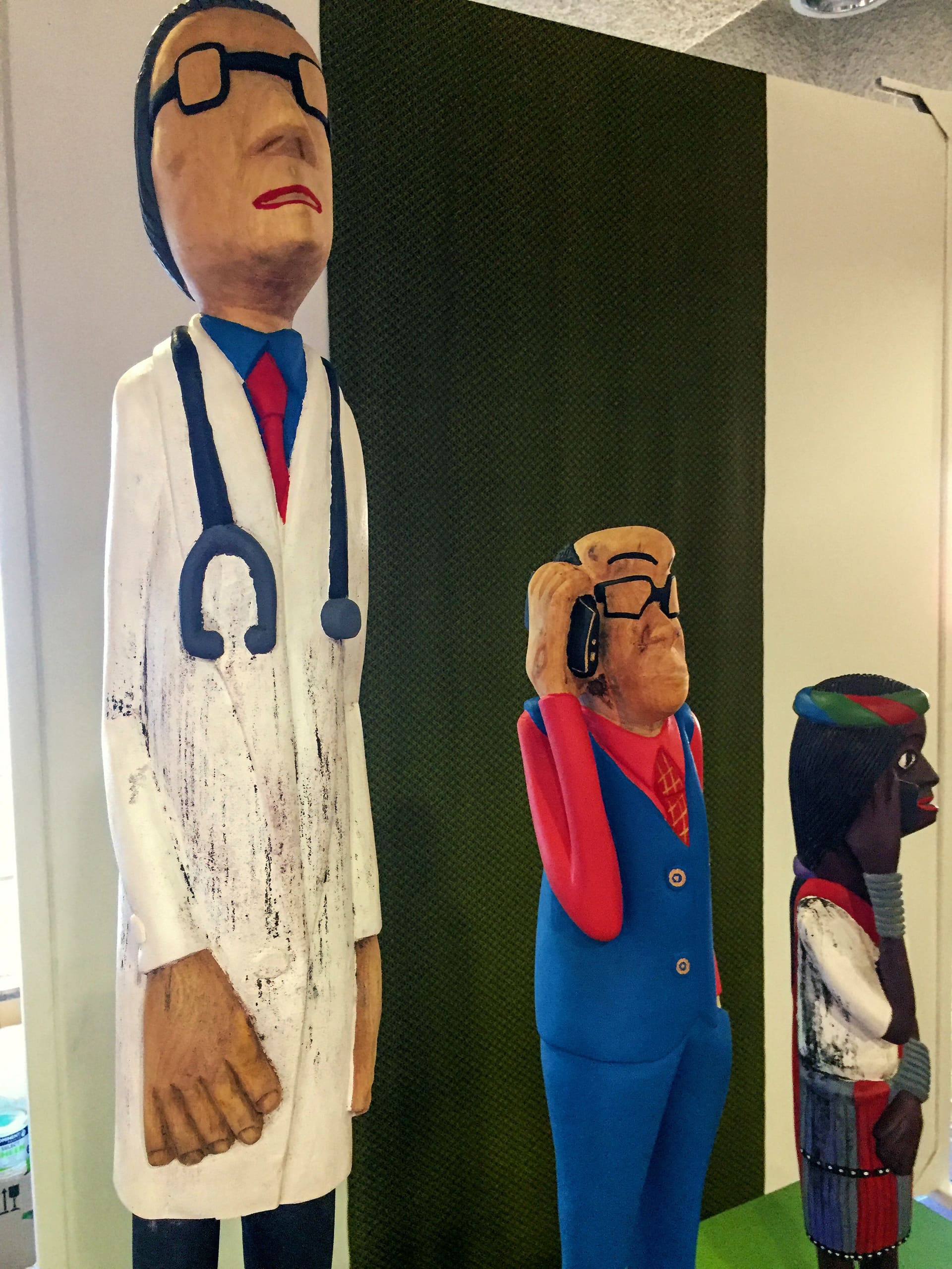 Three colourful carved wooden figures stand in a row. The tallest is a doctor with glasses, stethoscope, and white coat. The middle figure wears a blue suit and red tie, holding a phone to his ear. The smallest is dressed in vibrant, patterned clothes and a green cap.
