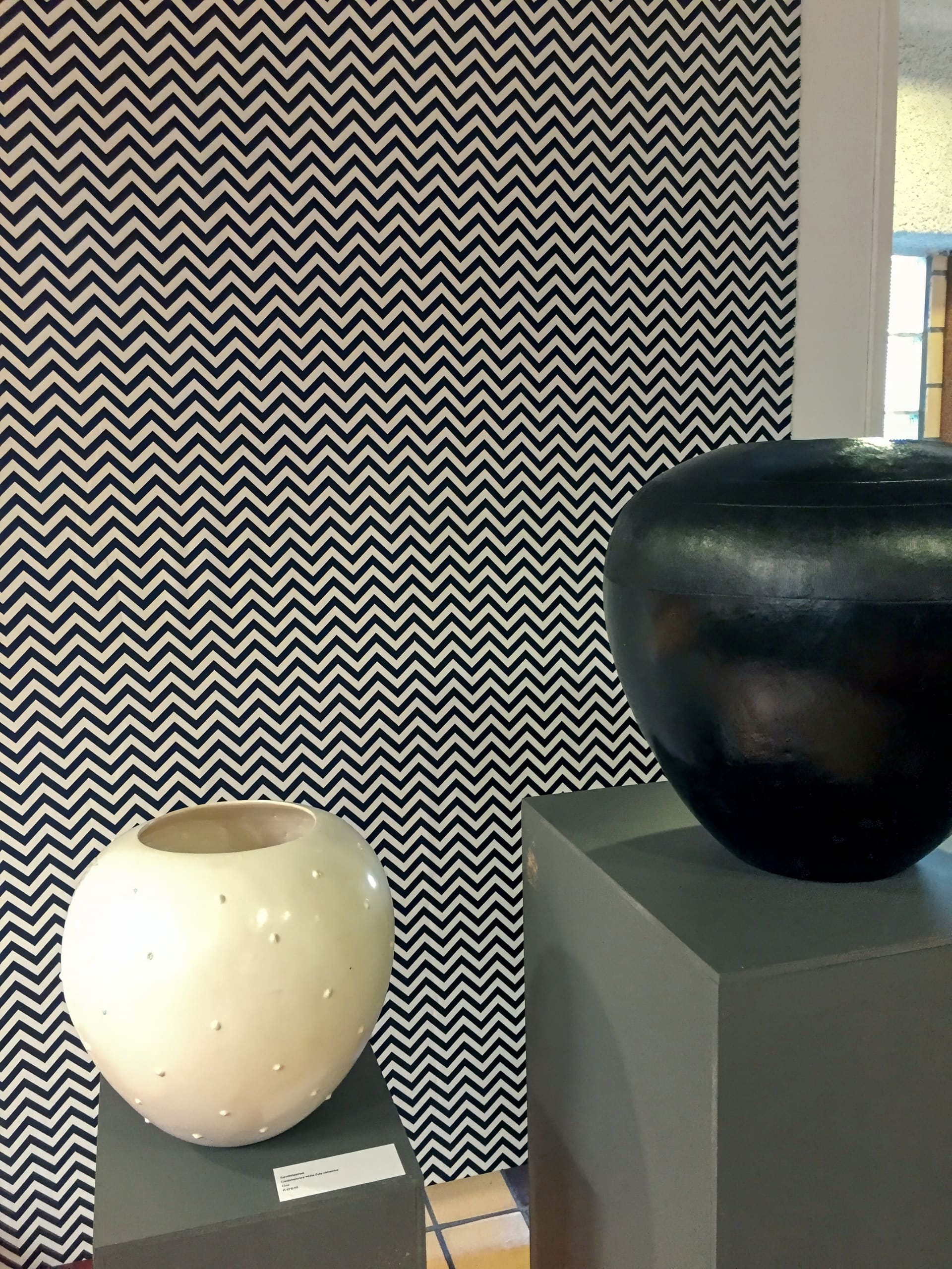 Two ceramic vases sit on grey pedestals; the left vase is round, white, and dotted with small holes, while the right vase is smooth, larger, and black. The background features bold black-and-white chevron stripes.