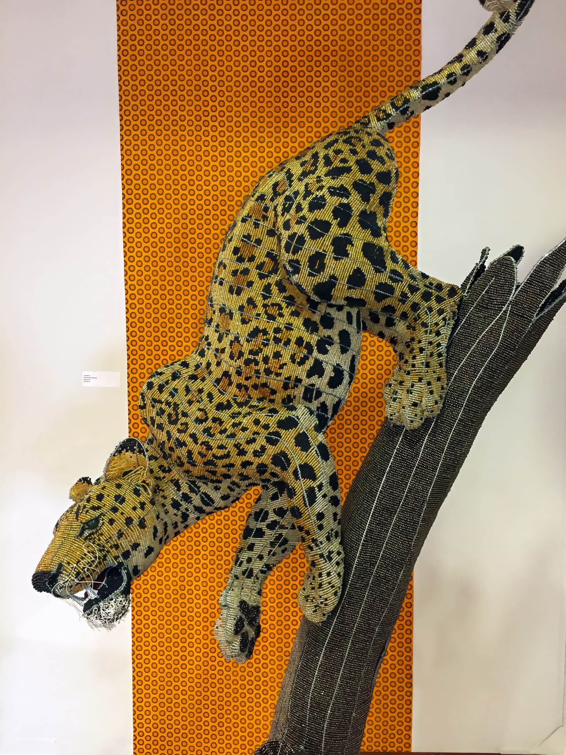 Large sculpture of a leopard made from beads climbs a tree branch against an orange background with a repeating black pattern. The leopard is yellow with black spots and intricate beadwork, giving a textured, lifelike appearance.