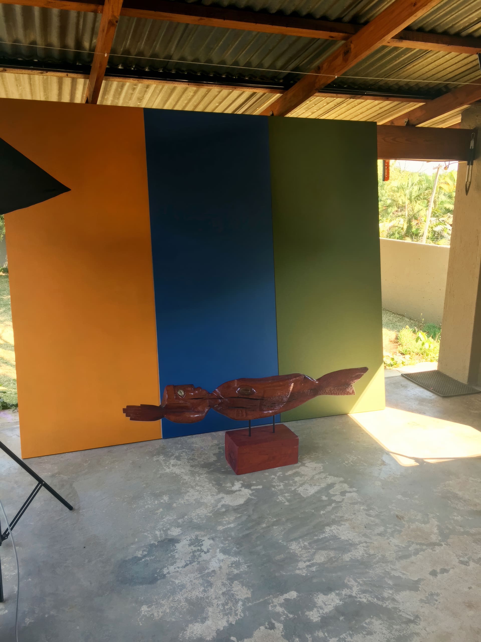 A wood sculpture of a fish on a red base sits on a concrete floor in front of a backdrop with vertical panels in orange, blue, and green. Sunlight filters in from the right, with part of a black studio light visible on the left.