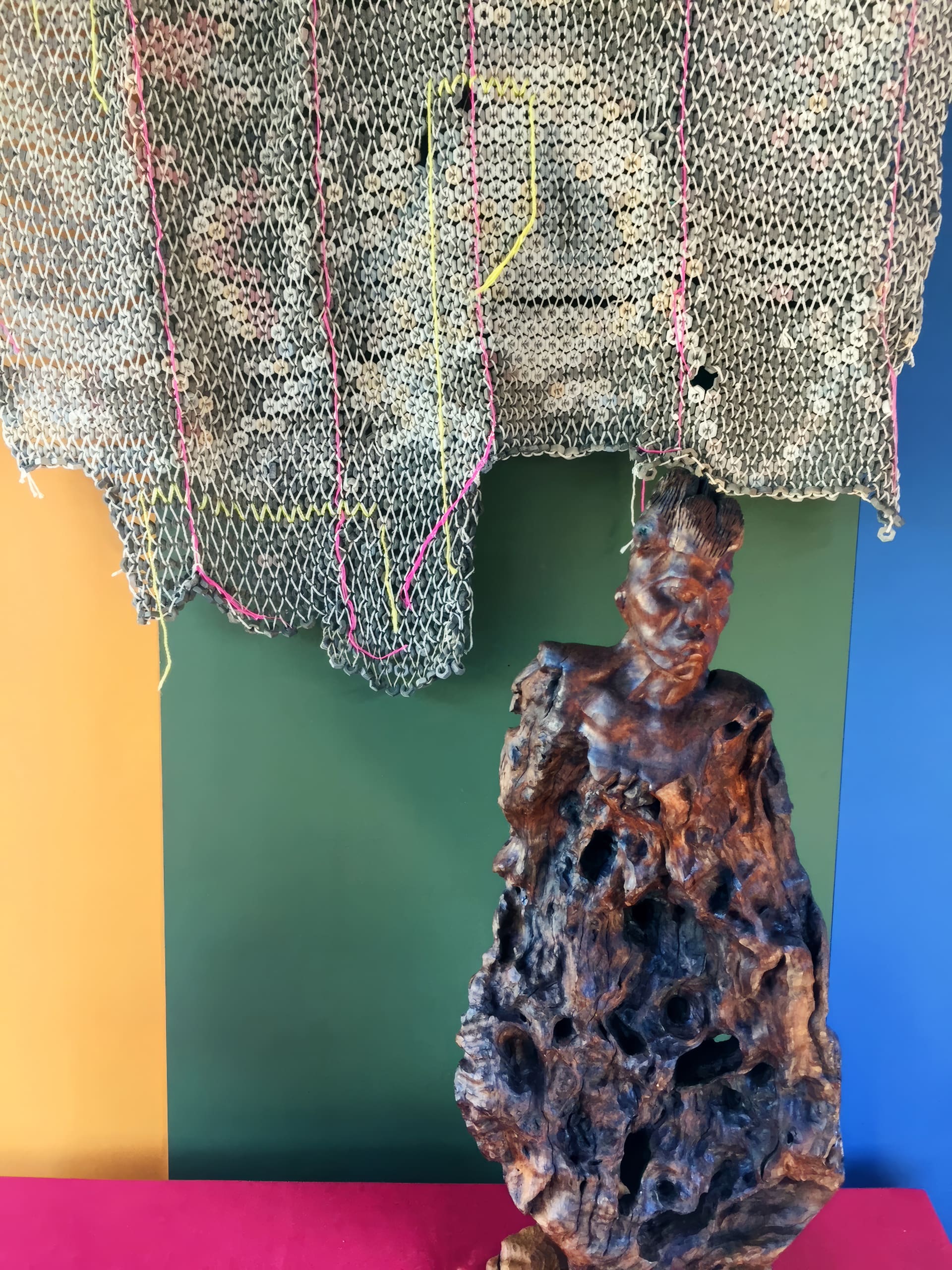 A carved wooden sculpture of a person is placed in front of a colour-blocked wall with yellow, green, and blue sections. Above the sculpture hangs a loosely woven, net-like textile with vertical coloured threads running through it.