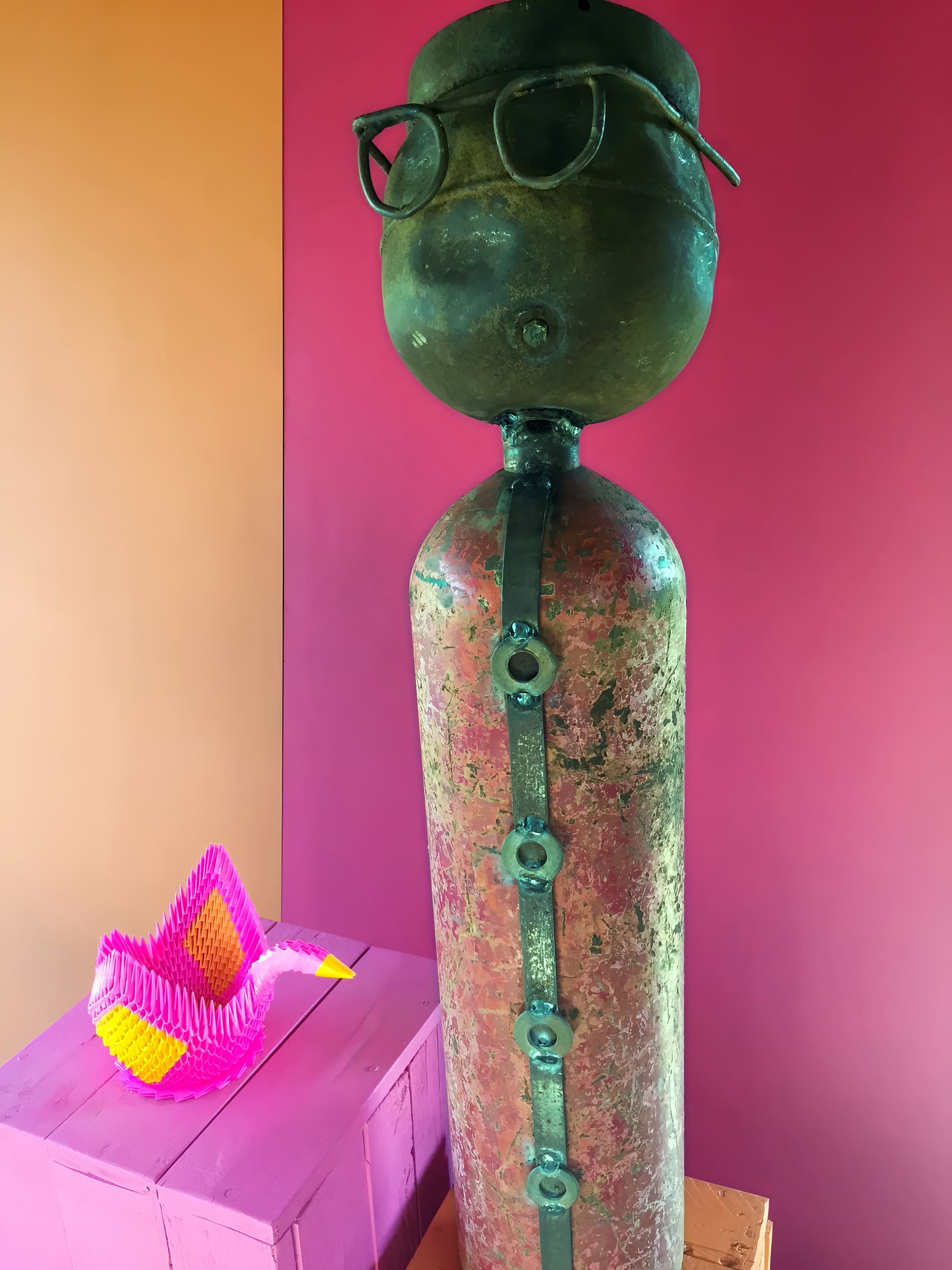 A tall, weathered metal cylinder sculpture with a round top, black-rimmed glasses, and bolt details stands against a pink wall. Nearby, a folded pink and yellow paper object rests on a purple box, with an orange wall in the background.