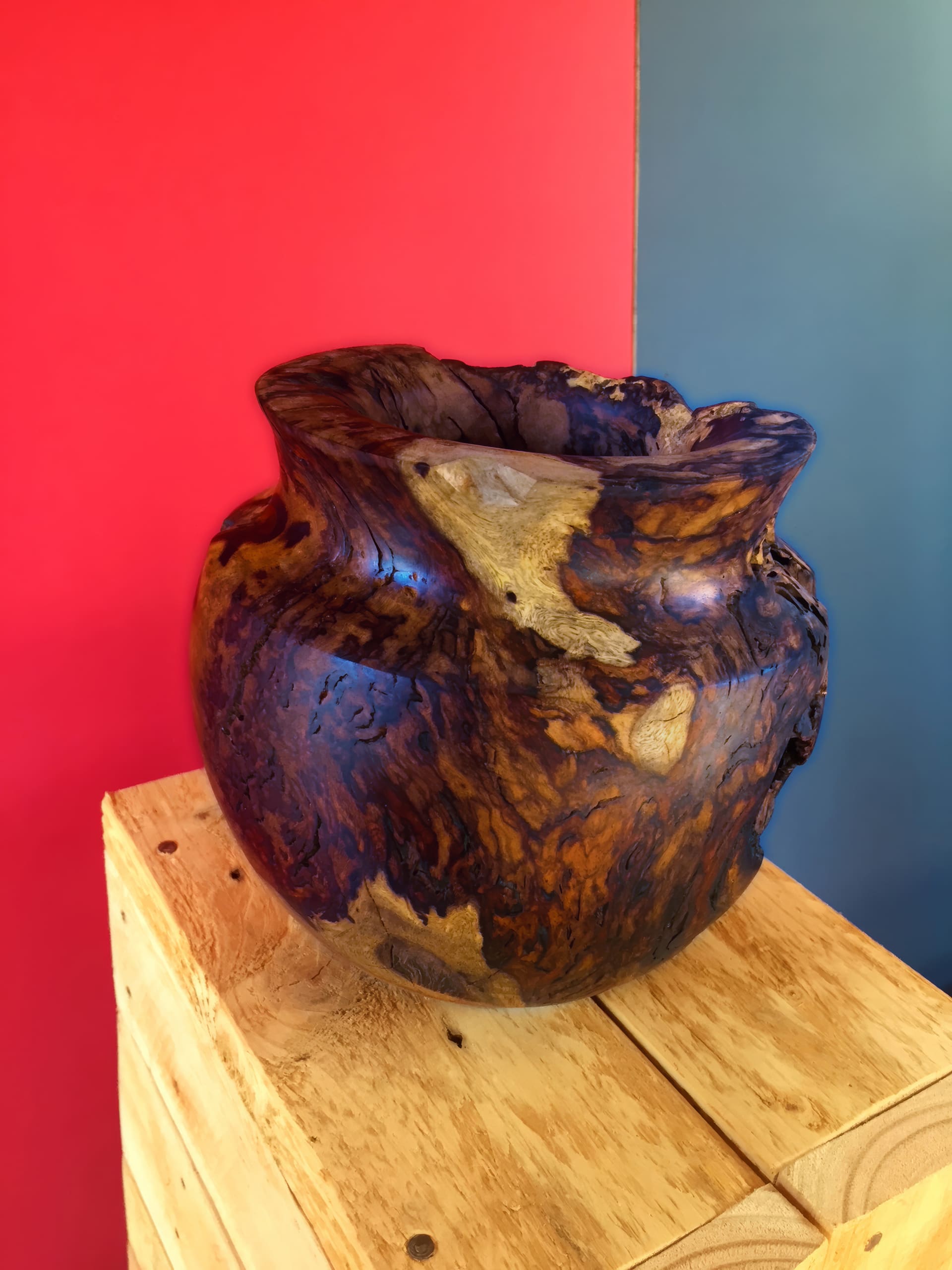 A rustic, round wooden vase with a rough, natural texture and varied brown tones sits on a light wooden box. The background is split vertically with a red section on the left and a blue-grey section on the right.