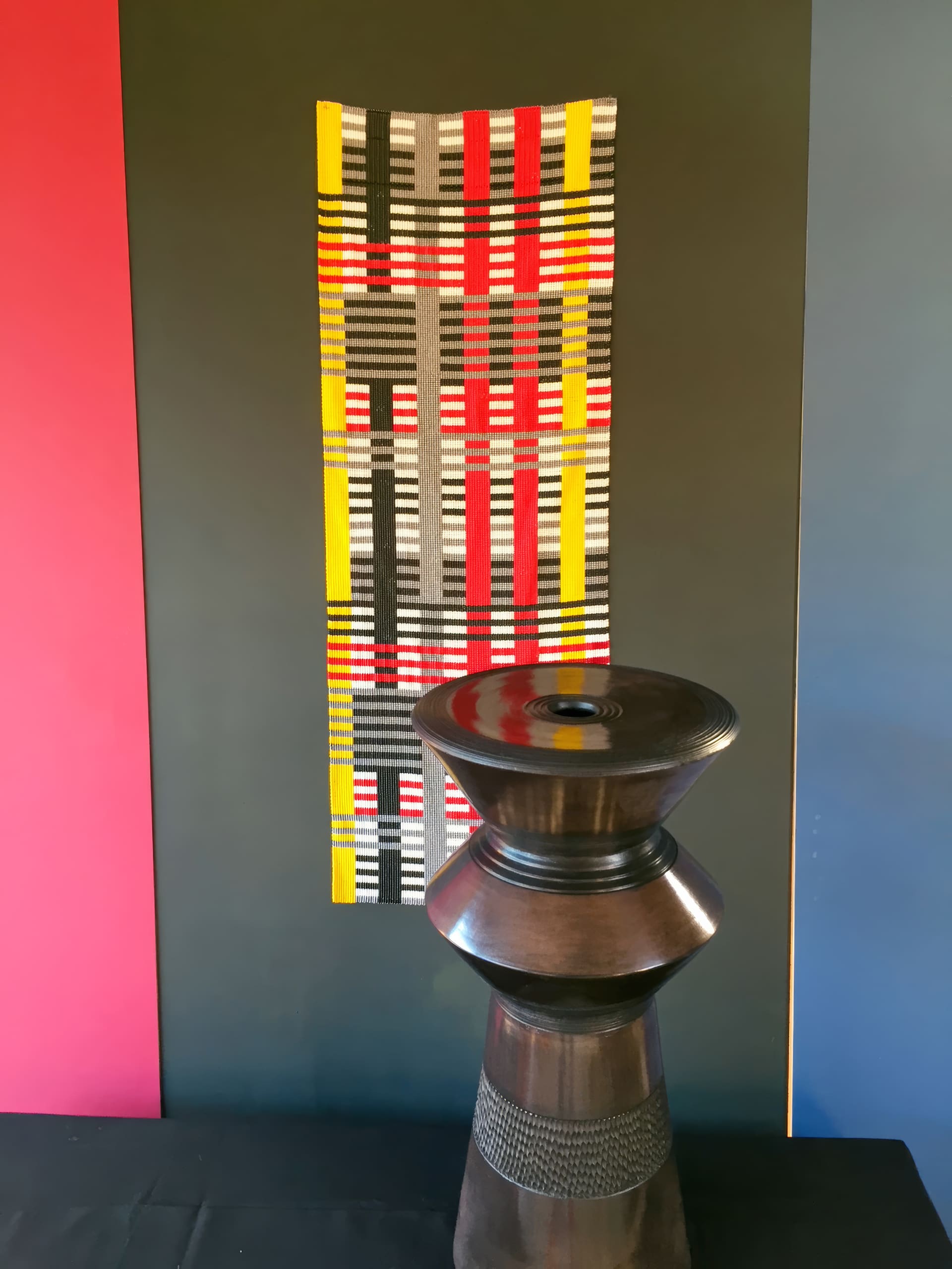 A dark wooden sculptural object stands on a black surface in front of a vertical textile with red, yellow, black, grey, and white horizontal and vertical stripes. The background has red, black, and blue colour blocks.