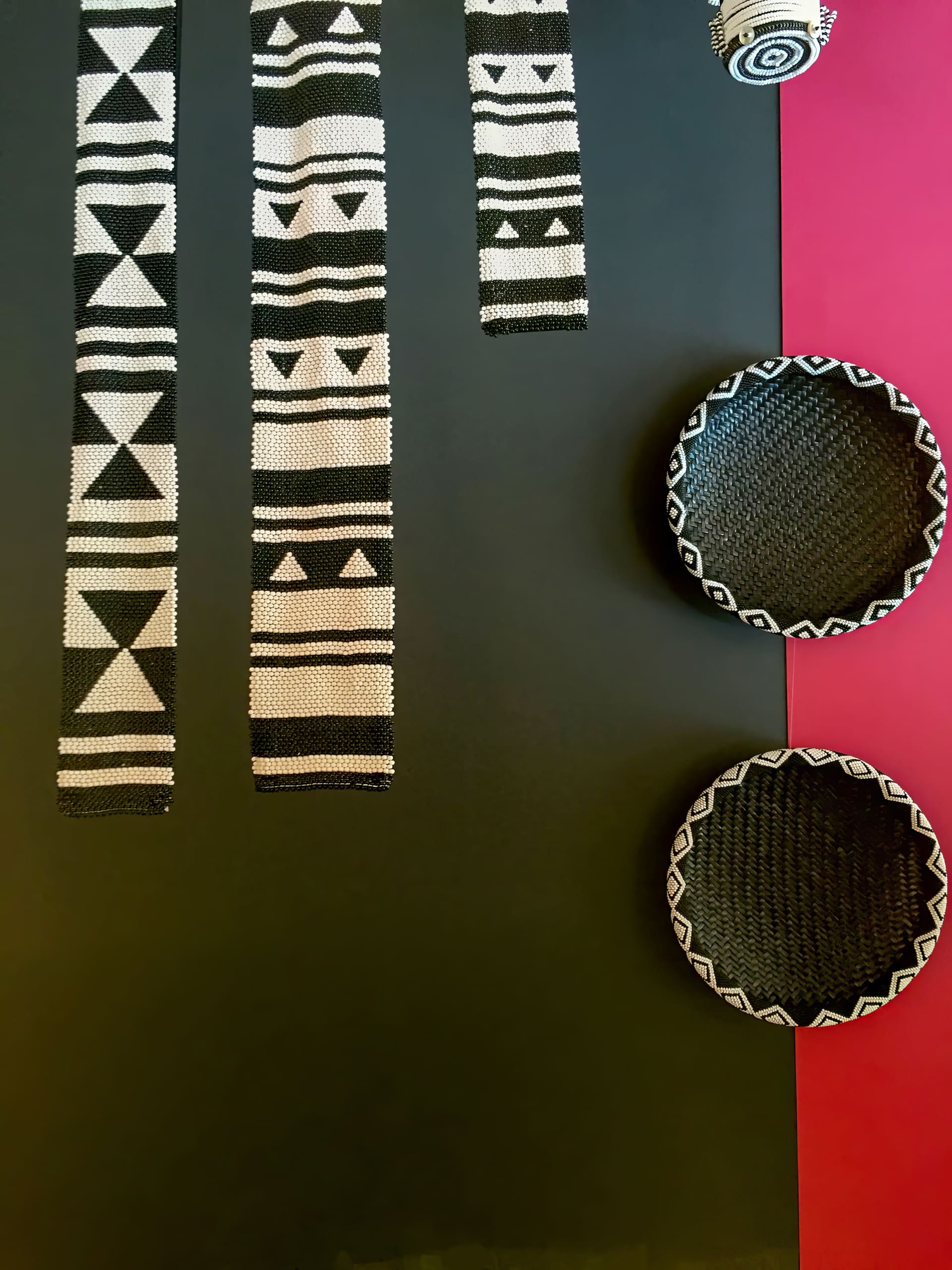 Three black and white baskets with geometric patterns hang on a wall split vertically: black on the left and red on the right. Four woven black and white bands featuring triangles and stripes hang vertically on the black section.