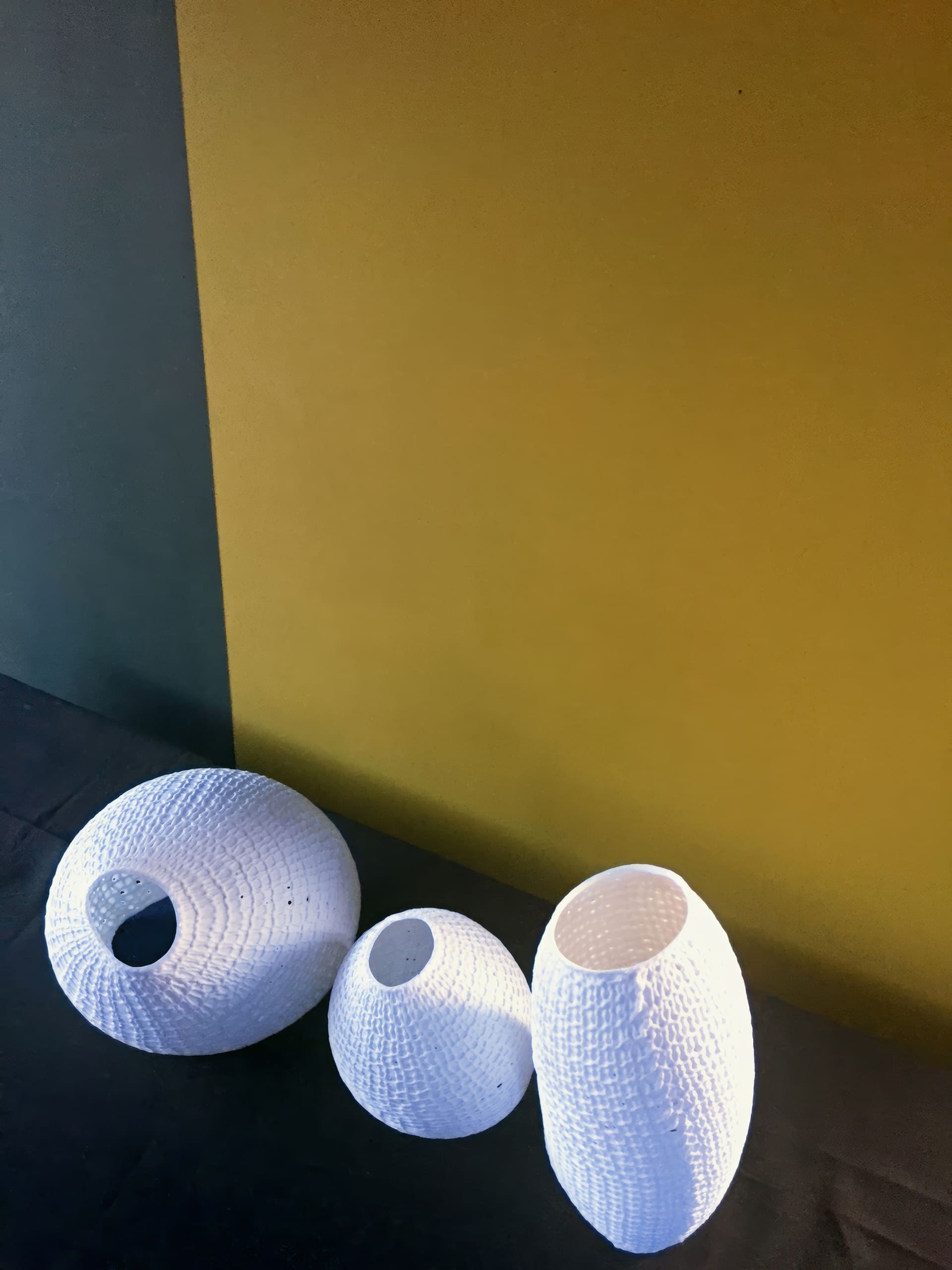 Three white, textured, oval vases of varying heights are arranged on a dark surface. The background is divided vertically, with the left side a muted green and the right side a mustard yellow. Bright light highlights the vases' patterns.