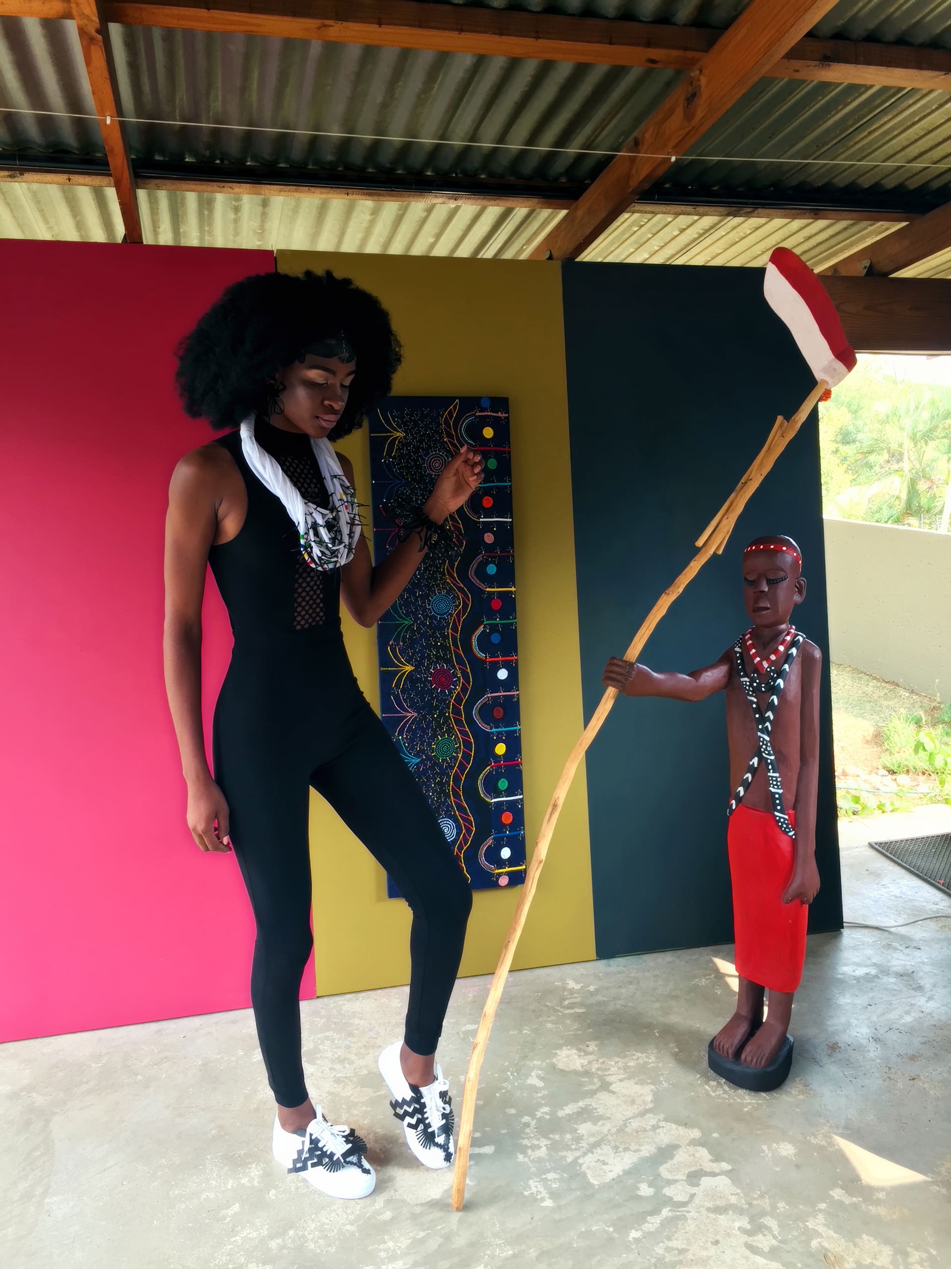 A tall person with a black afro wig, wearing a fitted black outfit, white scarf, and patterned trainers, stands next to a carved wooden statue of a person in red attire holding a tall staff. They pose in front of a colourful, patterned background panel.