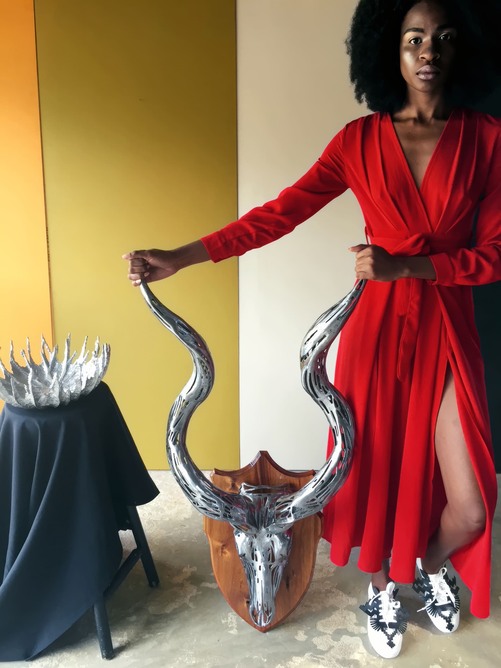 A woman in a long red dress and white trainers stands on a tan floor, holding a mounted silver antelope skull with curved horns. Beside her is a black-draped table displaying a shiny, metallic crown. The wall behind her is divided into gold and cream sections.