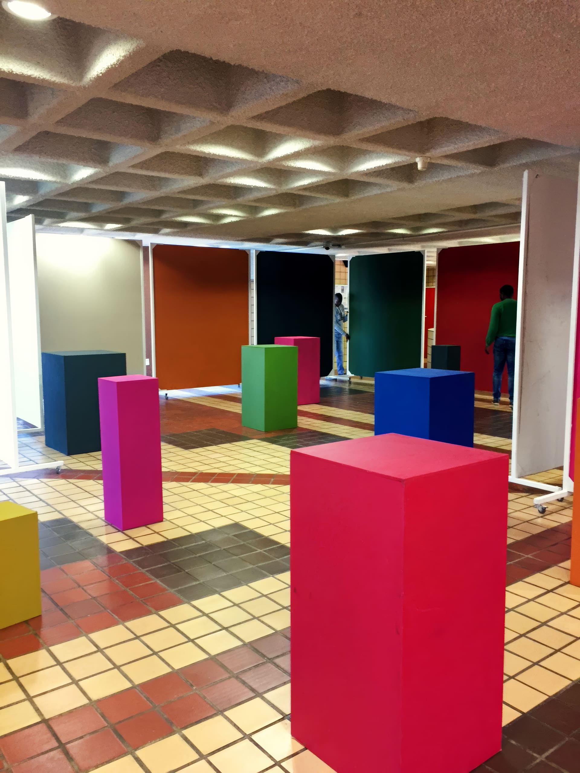 An indoor space with several colourful rectangular plinths—pink, yellow, blue, green—on a tiled floor. The background features large panels in red, white, green, orange, and grey. Two people stand near the back. The ceiling has a grid pattern.