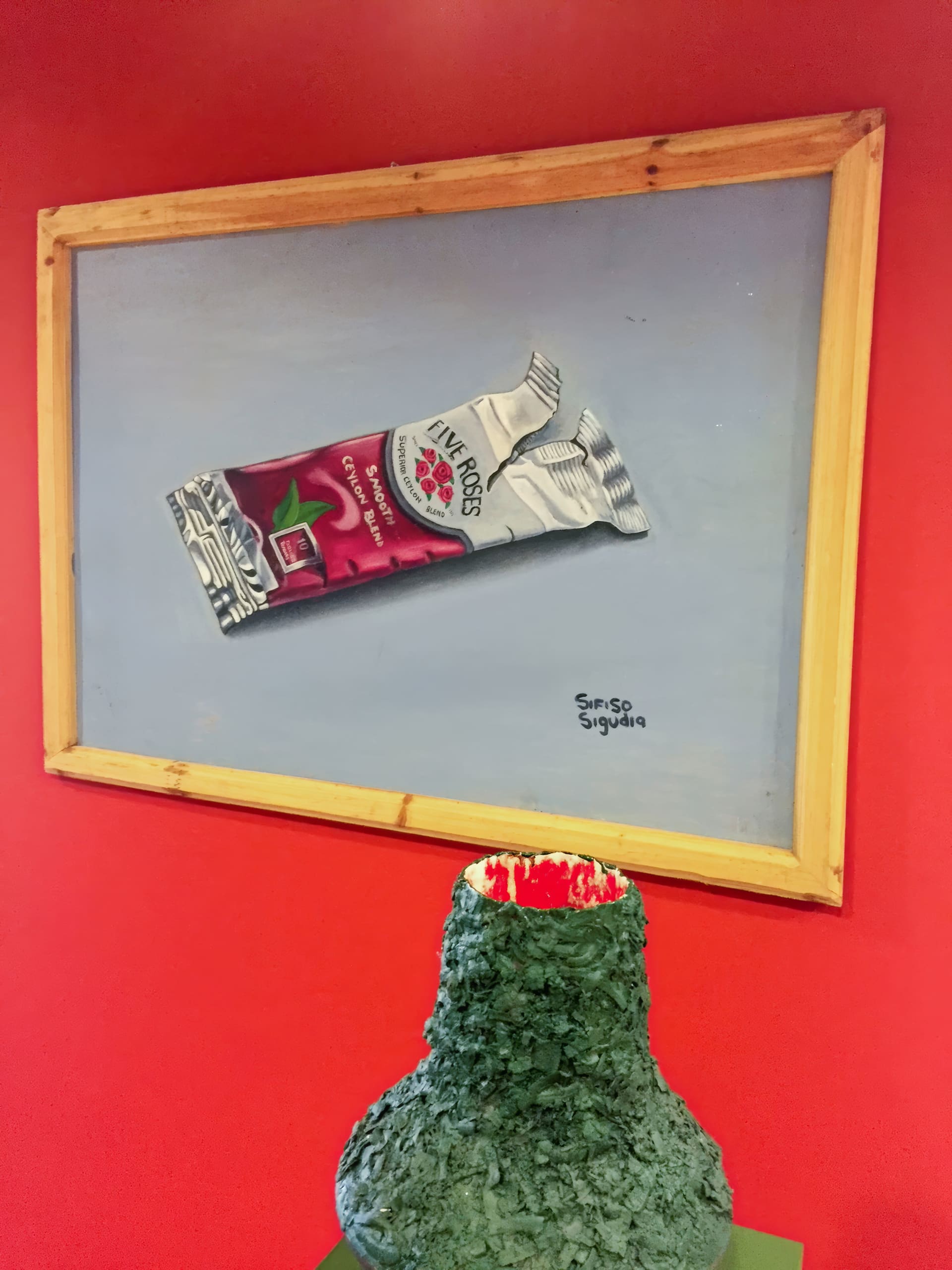 A framed painting of a Five Roses cranberry & apple tea packet hangs on a bright red wall above a green, volcano-shaped sculpture with a red, lava-like top. The artist’s signature, “Simiso Soyiya”, appears on the painting.