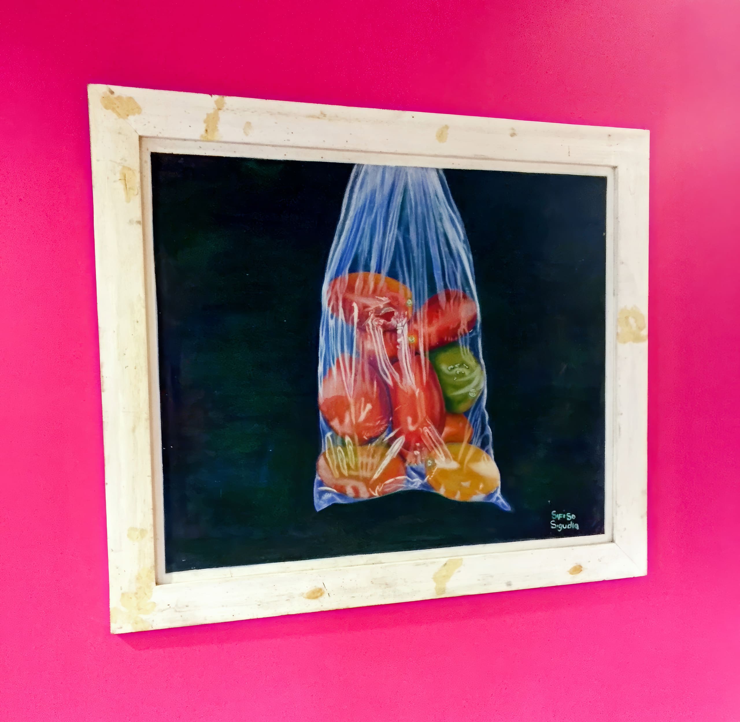 A framed painting hangs on a bright pink wall, depicting red, orange, and green peppers inside a transparent plastic bag, set against a dark background. The white frame shows spots of wear, and the artist's signature appears in the lower right corner.