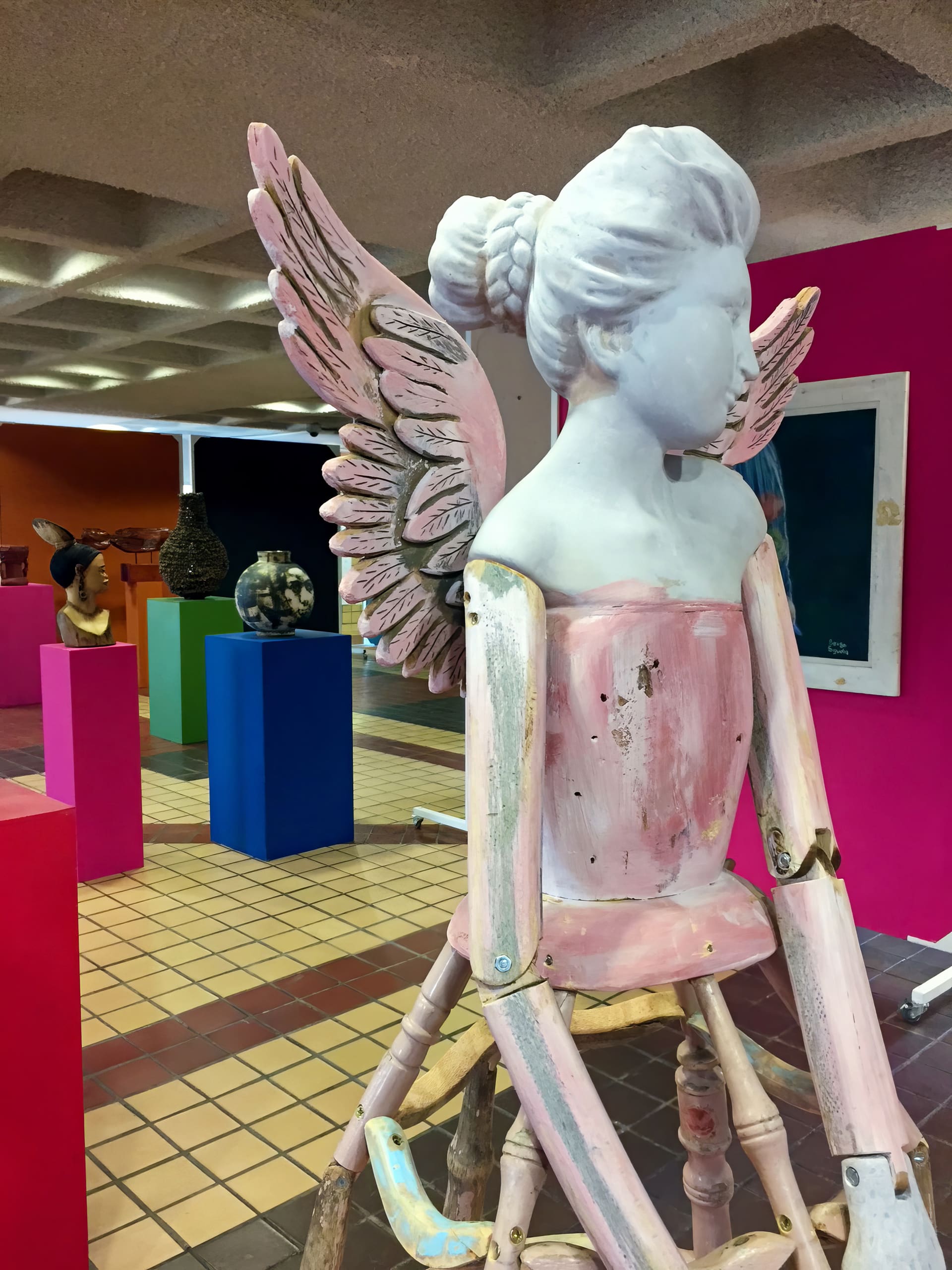 A wooden sculpture of a seated figure with plaited hair and pink angel wings is displayed in an art gallery with colourful plinths, tiled floors, and other artworks visible in the background. The figure's face and chest are painted white.
