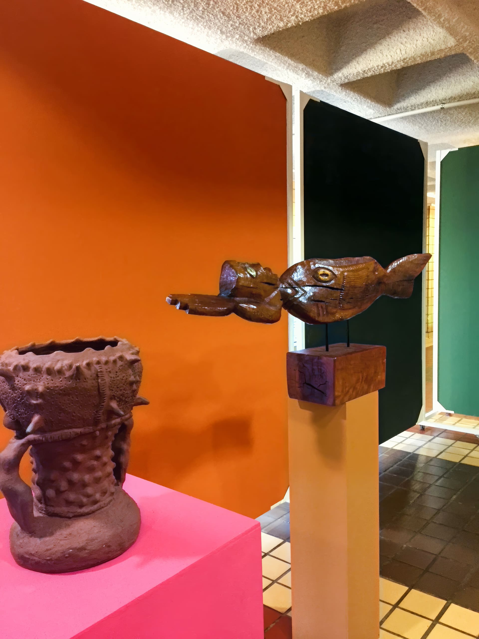 A textured brown ceramic vessel with handles sits on a pink plinth beside a wooden fish sculpture on a yellow stand. The background features large orange and dark green panels, and the floor is patterned with brown and cream tiles.