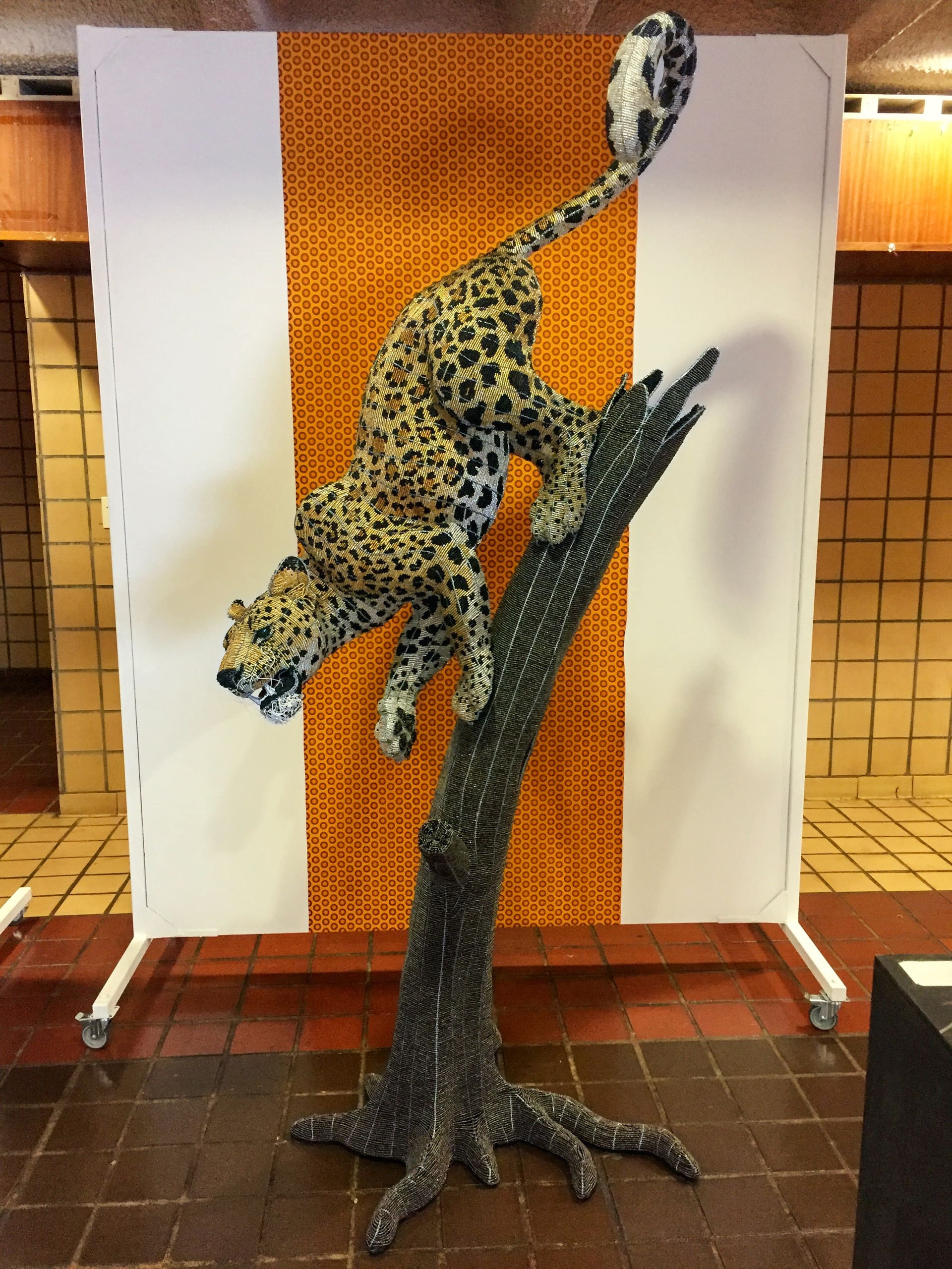 A realistic sculpture of a leopard leaps down from a tall, leafless tree branch. The background features a vertical orange panel with a honeycomb pattern, framed by cream tiles and brown floor tiles. The scene is indoors, with warm lighting.