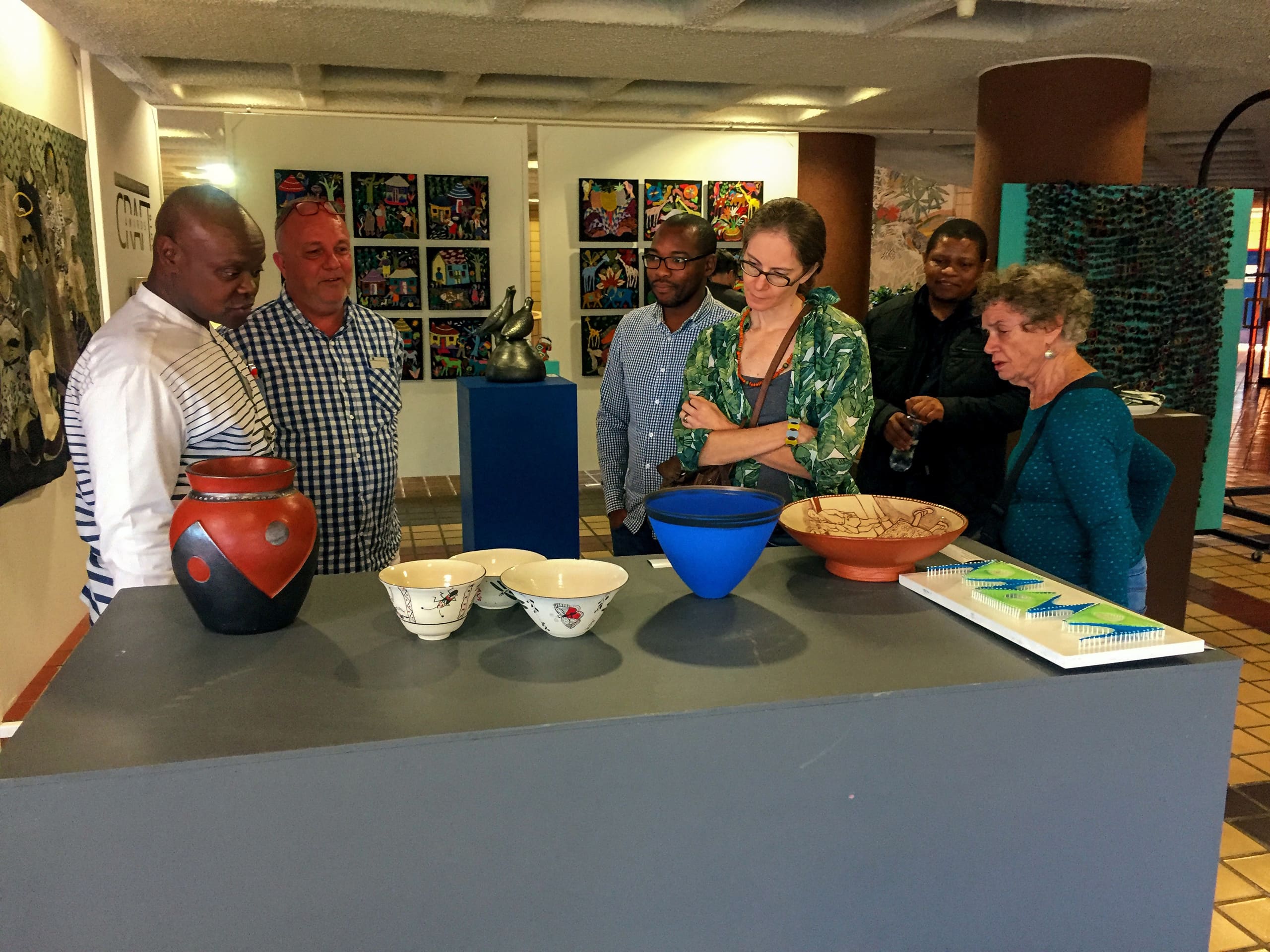 Six adults stand around a grey display plinth observing ceramic bowls and vases in an art gallery. Colourful abstract paintings hang on the walls behind them. The group appears engaged and thoughtful, with sketchbooks and artworks visible on the table.