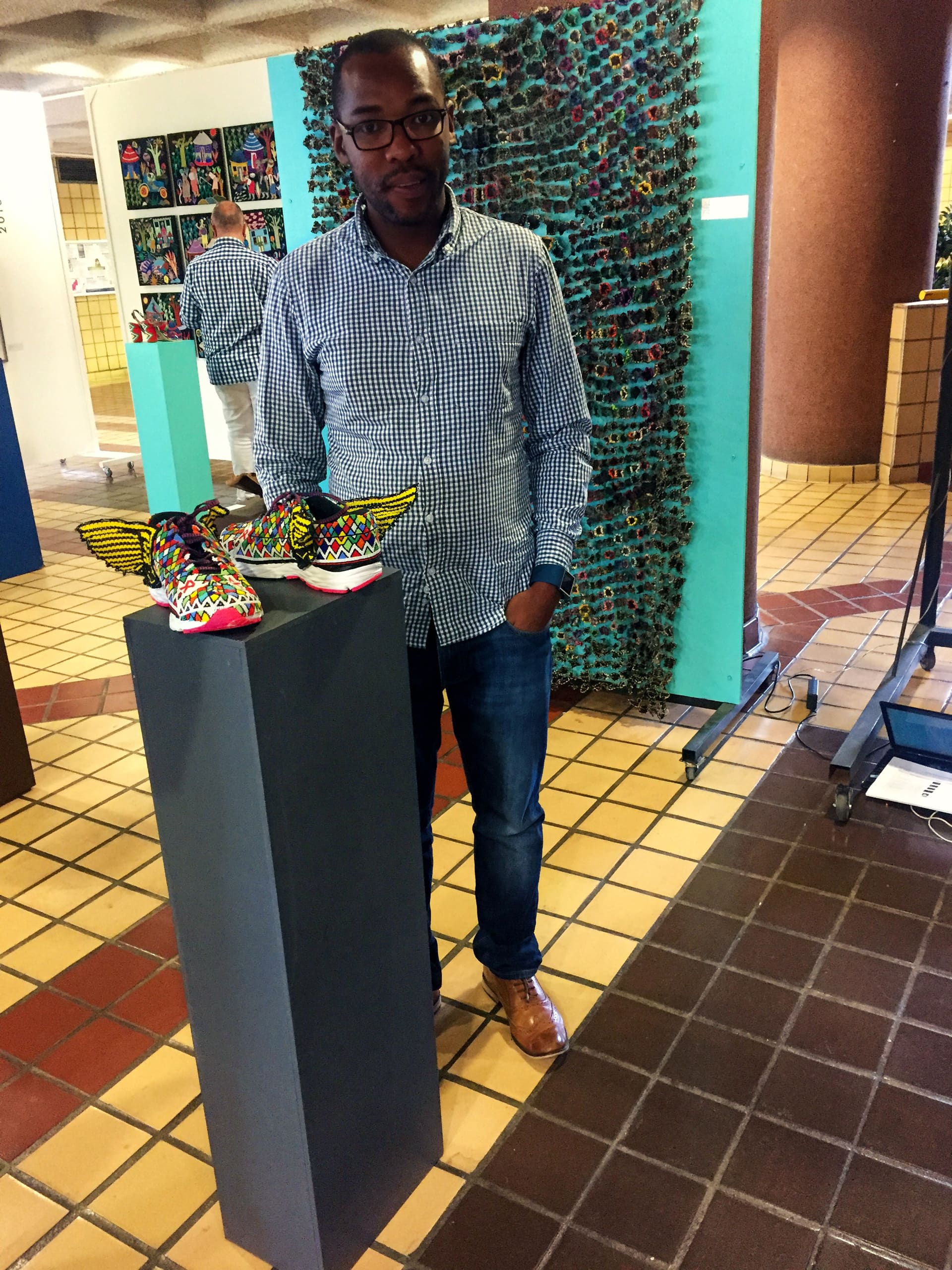 A man in glasses, a checked shirt, jeans, and brown shoes stands indoors beside a grey pedestal displaying colourful, winged shoes. Behind him are art panels covered in vibrant, small items, and the floor is tiled in beige and brown.